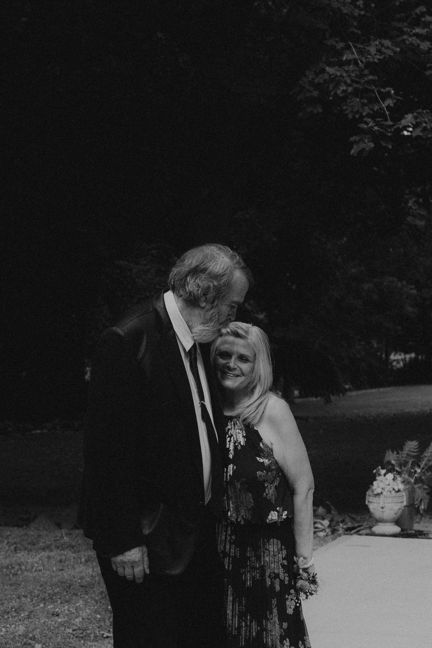 Black and white photo of a couple sharing a tender moment outdoors at a formal event, man in suit kissing womans head.
