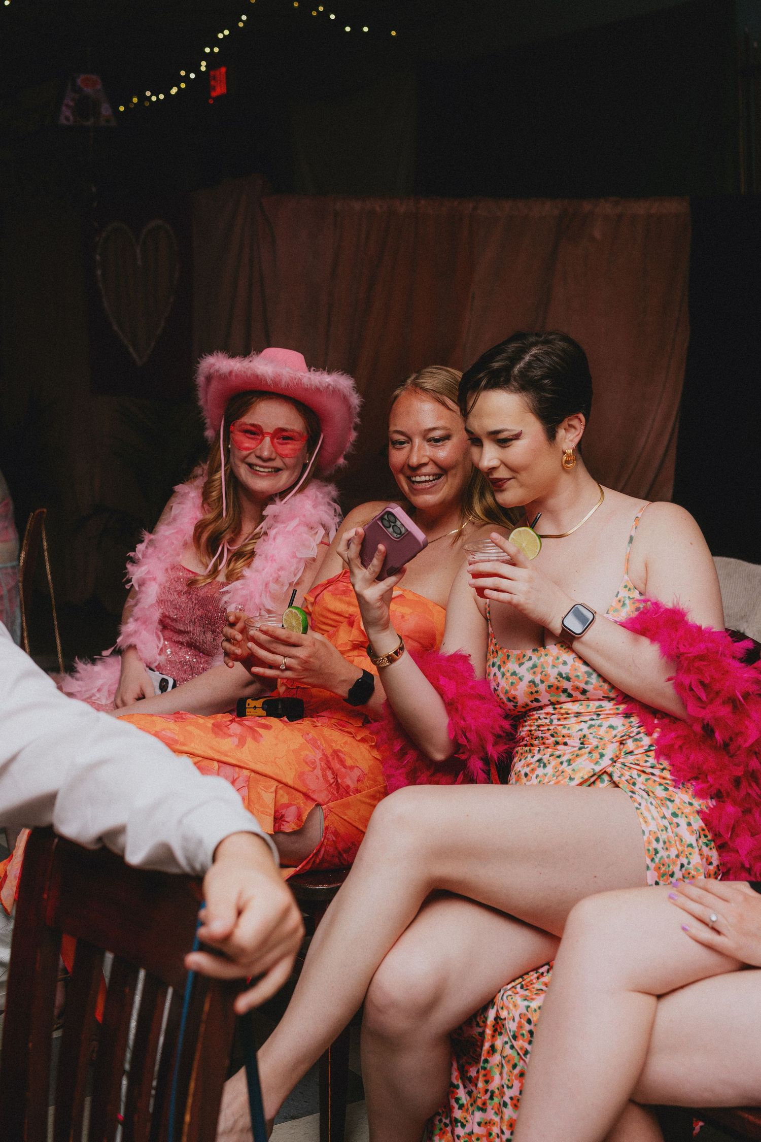Three women in colorful party outfits with pink feather boas laugh while looking at a phone at a festive gathering.
