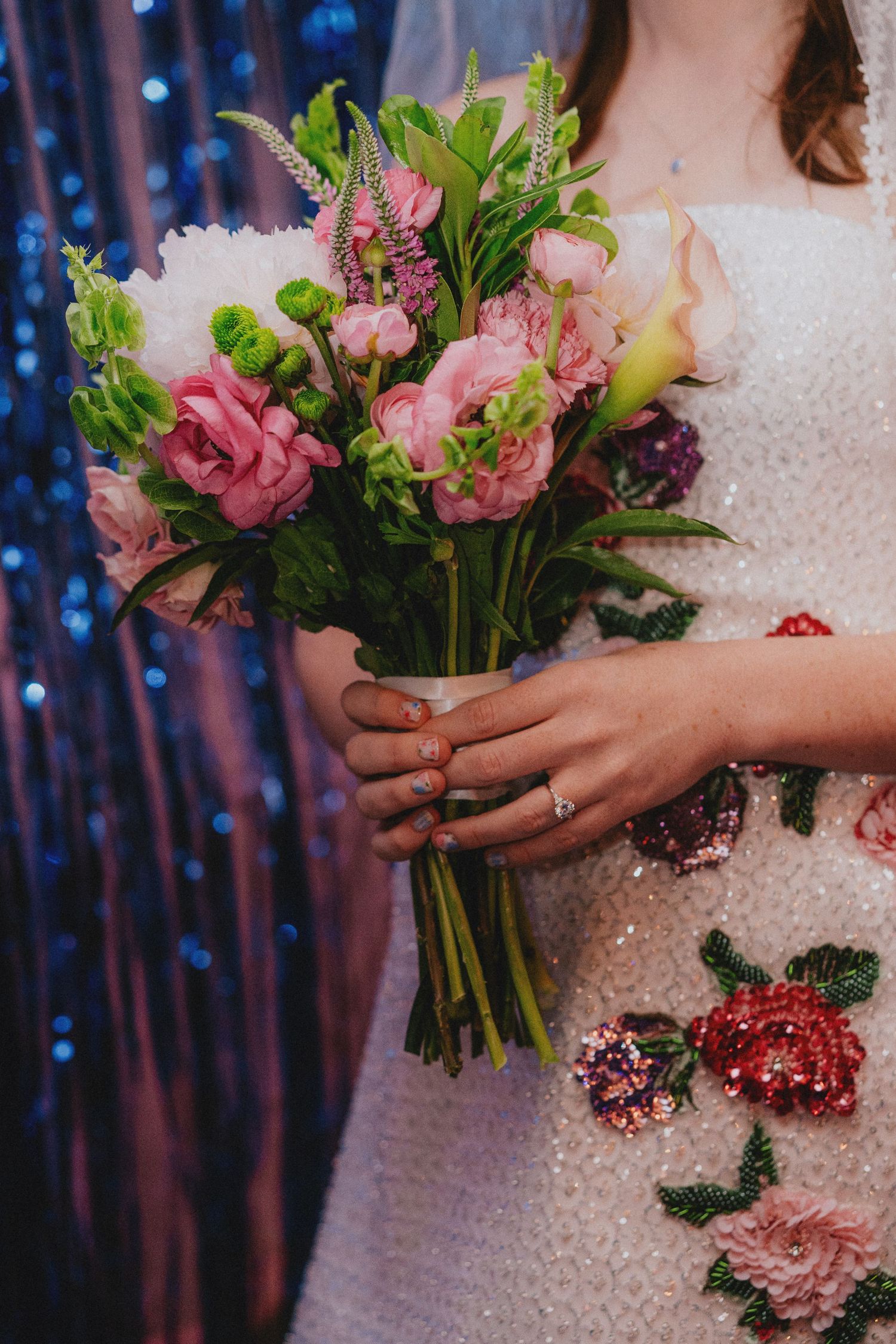 Bride in embroidered white gown holding pink floral bouquet with green accents against blue sequin backdrop.