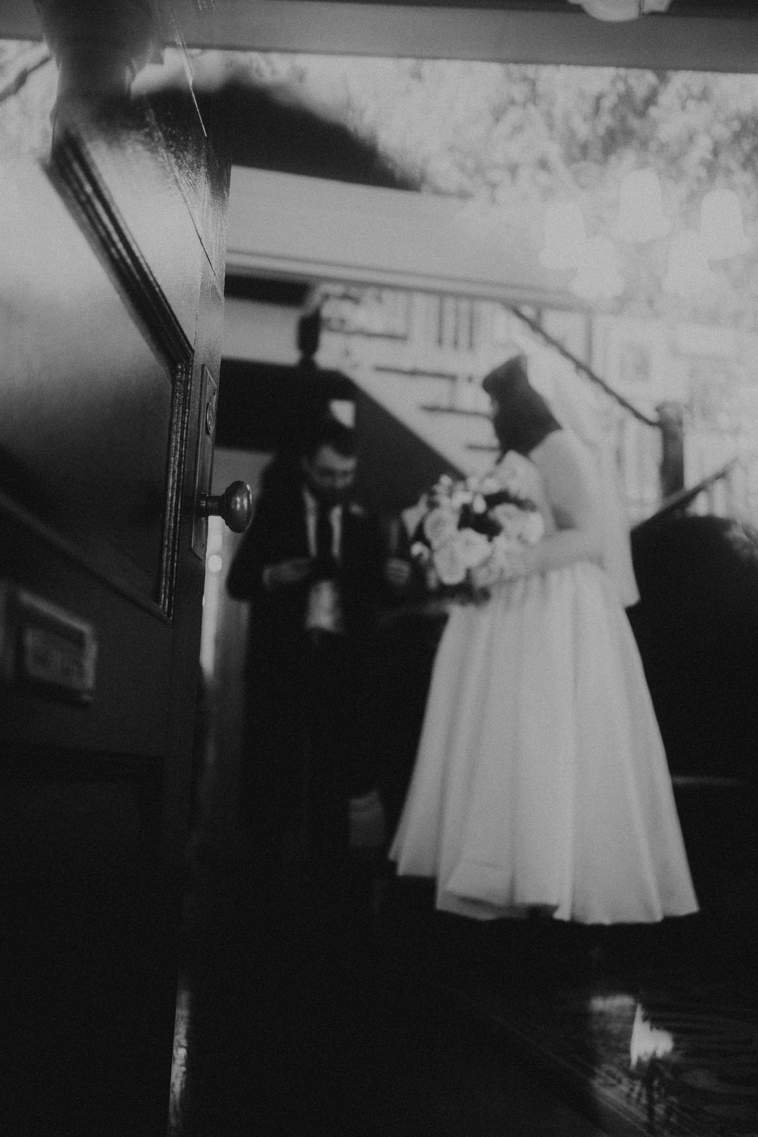 Black and white photo of a bride in white dress holding bouquet with groom in suit at a wedding venue entrance.