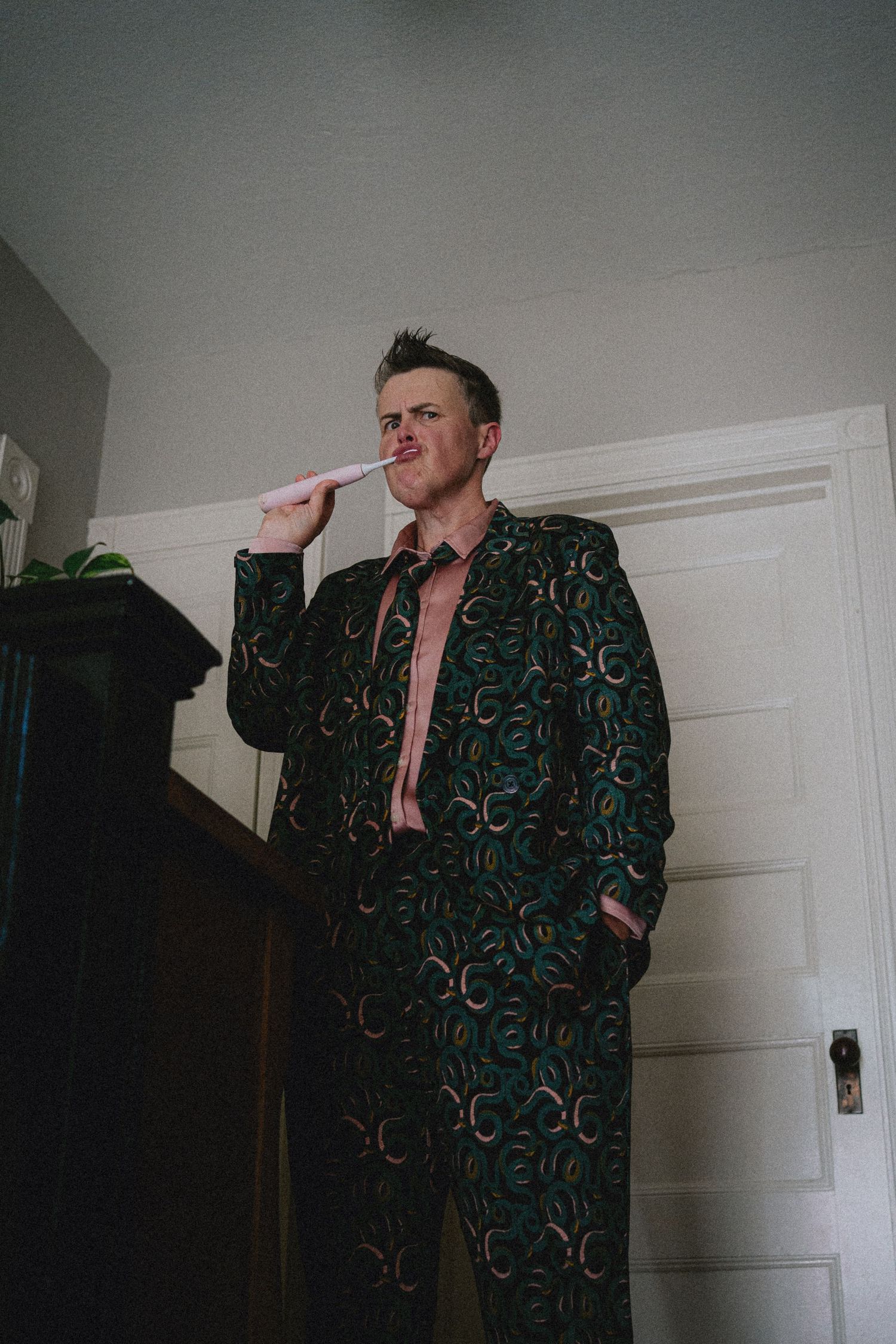 Man in bold green patterned suit holding a pen to his mouth, standing confidently indoors near a white door.