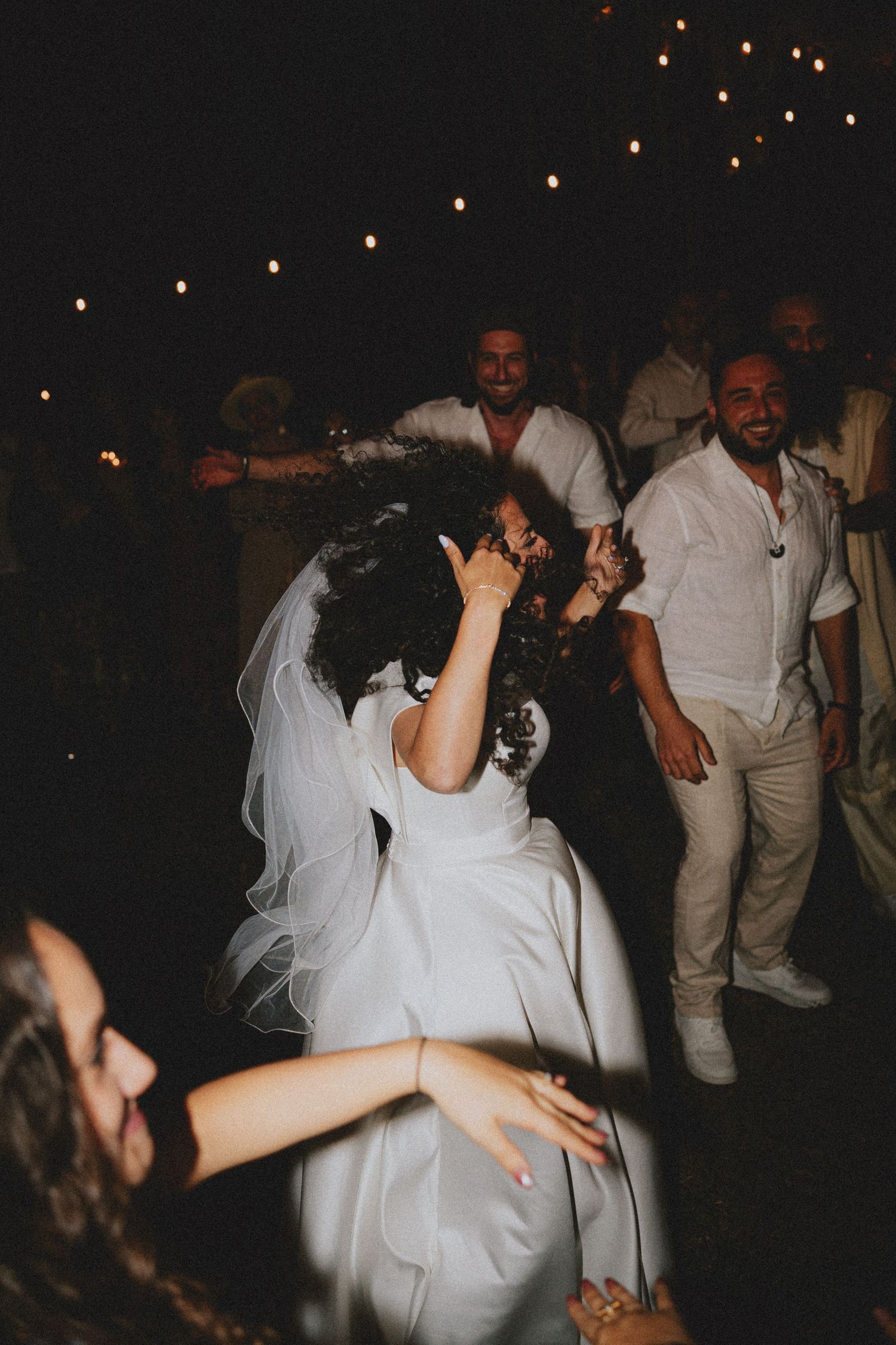 Bride in white gown and veil dancing joyfully with guests at an outdoor evening wedding reception.