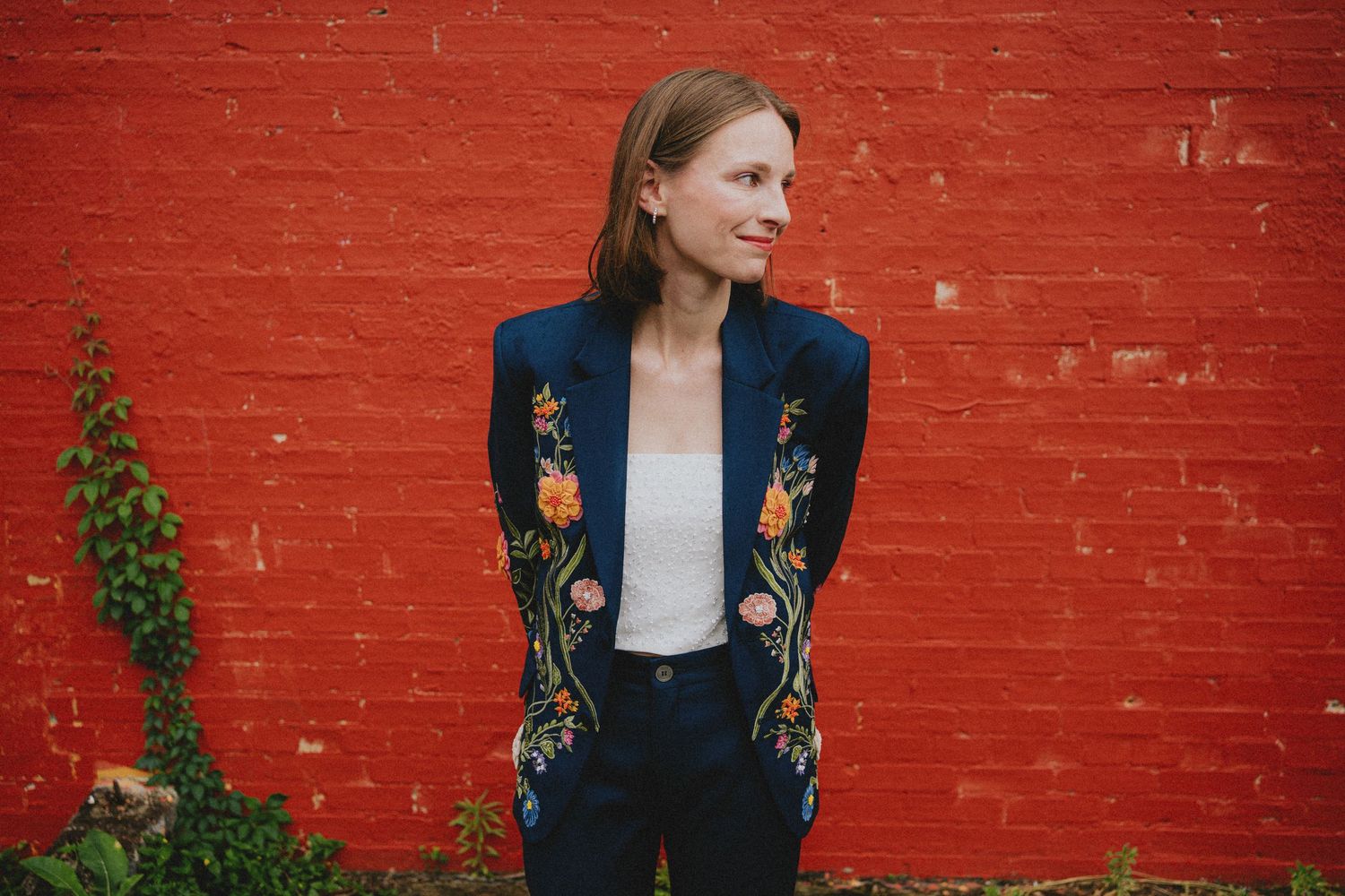 Woman in embroidered navy blazer posing against a vibrant red brick wall outdoors.