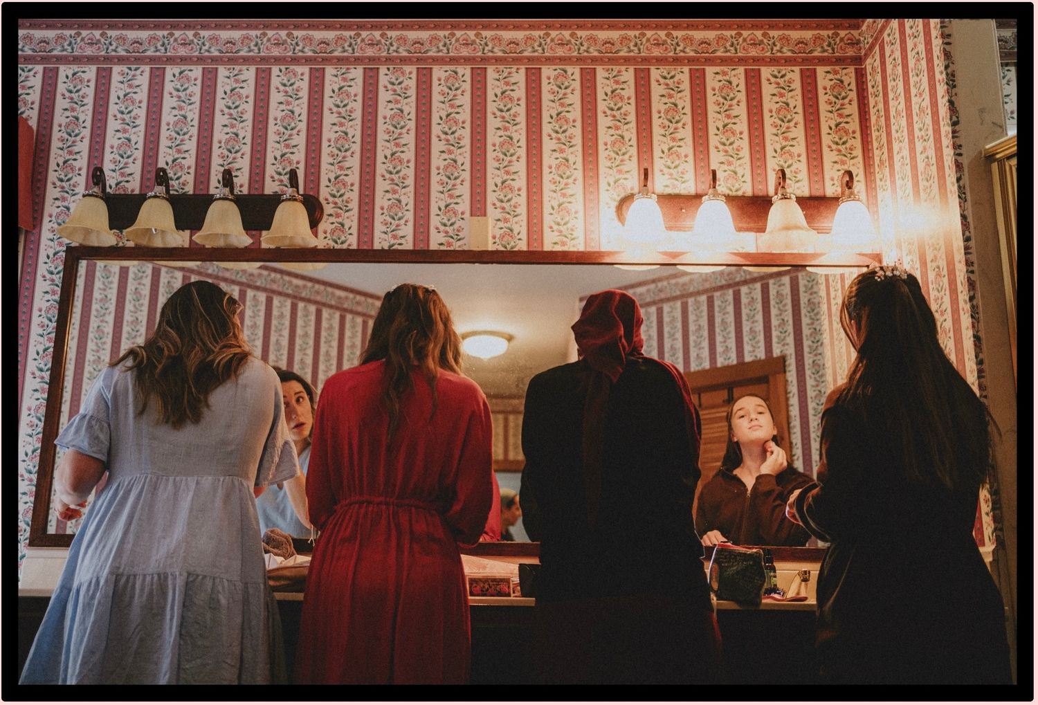 Four people stand with backs turned at a vintage diner counter with ornate wallpaper and warm pendant lighting.