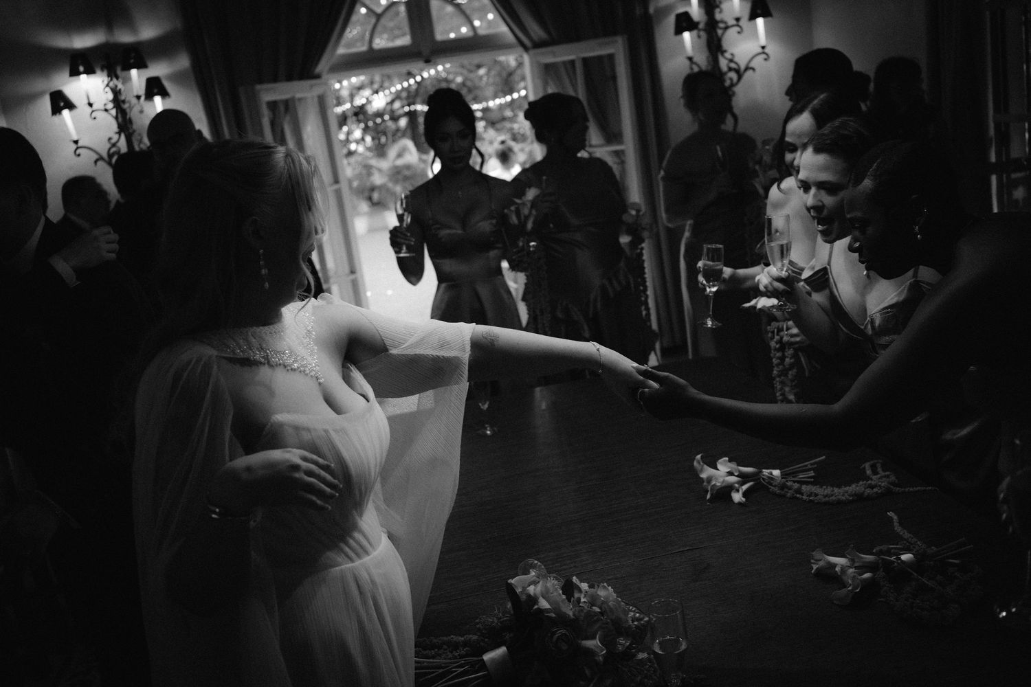 Black and white wedding photo of bride dancing and reaching out to guests at elegant indoor reception.