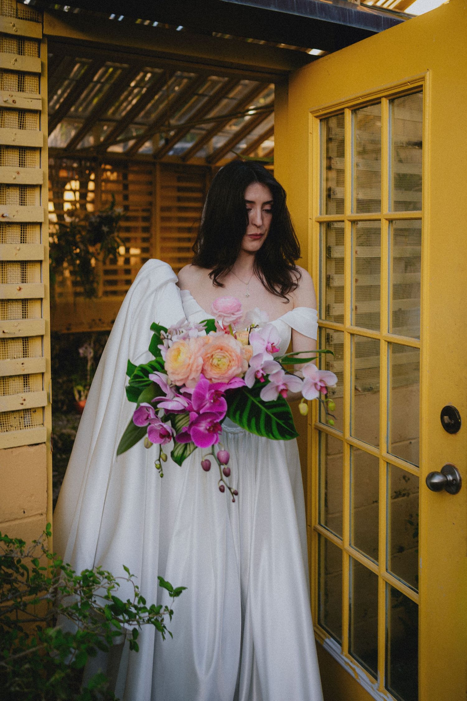 Bride in white gown holding vibrant pink orchid and peony bouquet standing by yellow French doors.