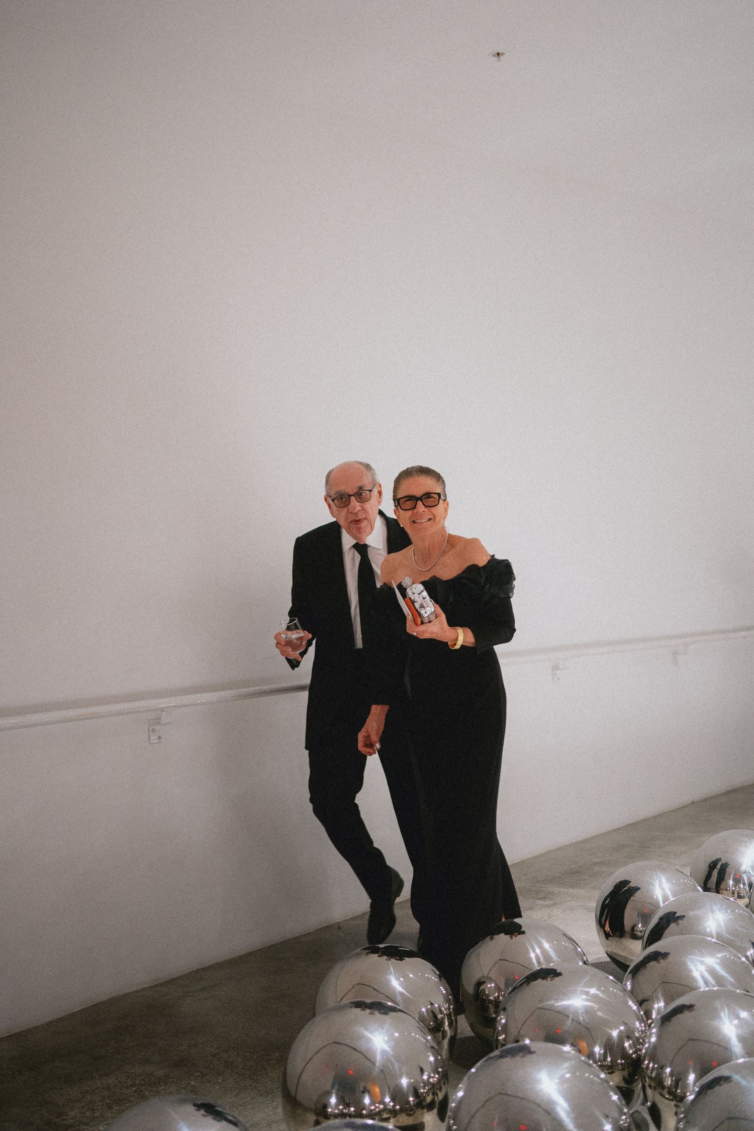 Elegant couple in black formal attire posing with silver mirror balloons at a chic gallery event.