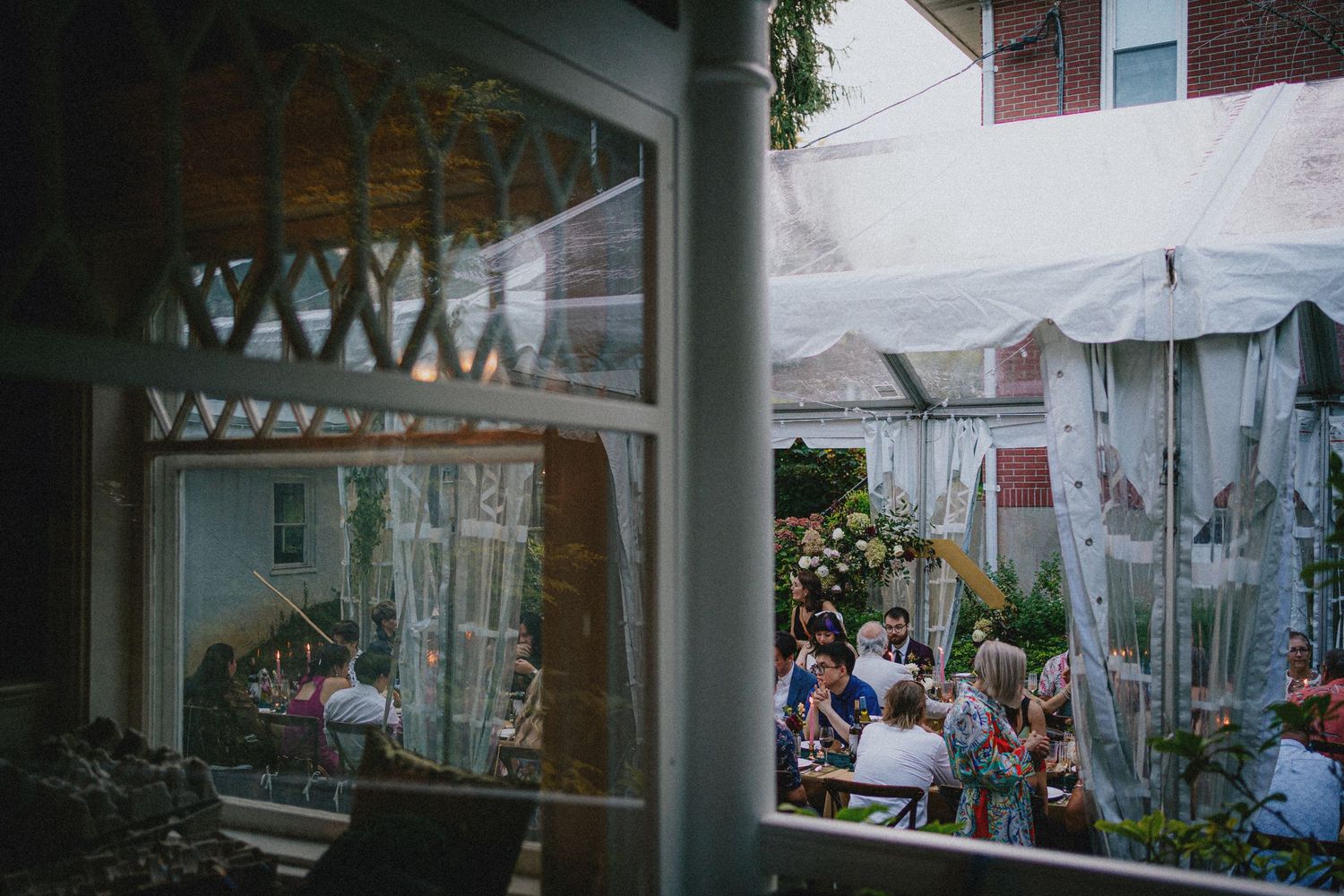 Outdoor garden party with guests gathered under a white tent beside a brick building with large windows.