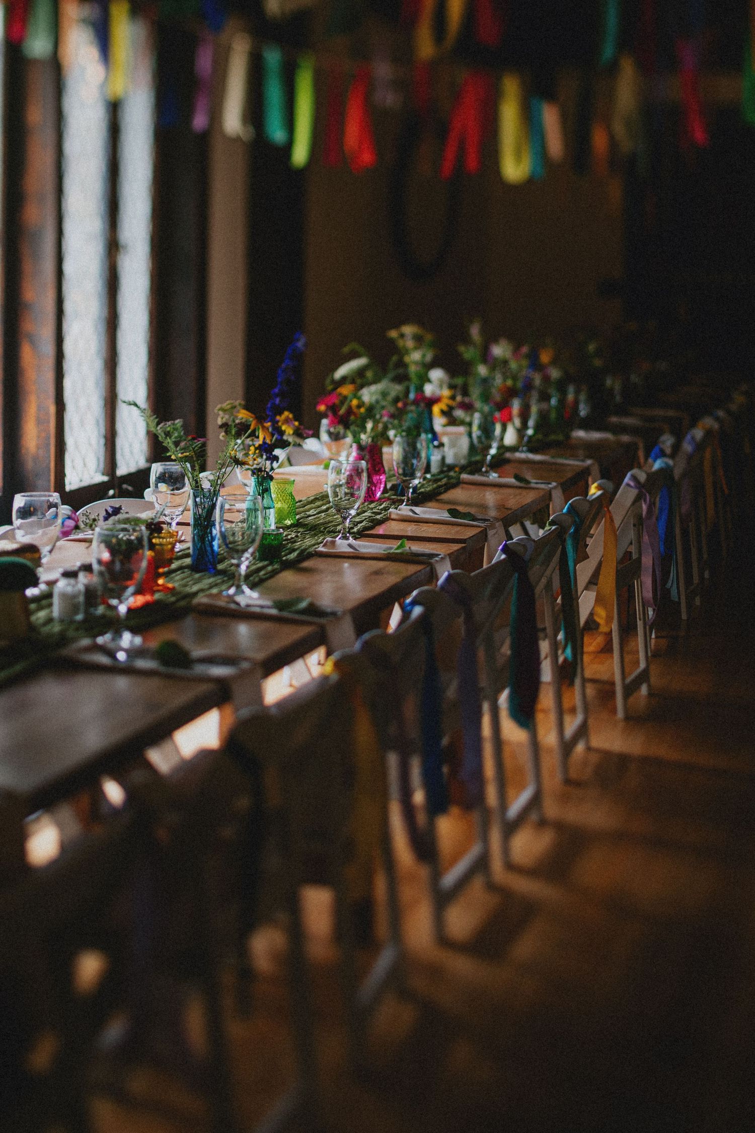 Colorful wedding reception table with floral centerpieces, rainbow ribbons on chairs, and festive bunting overhead.