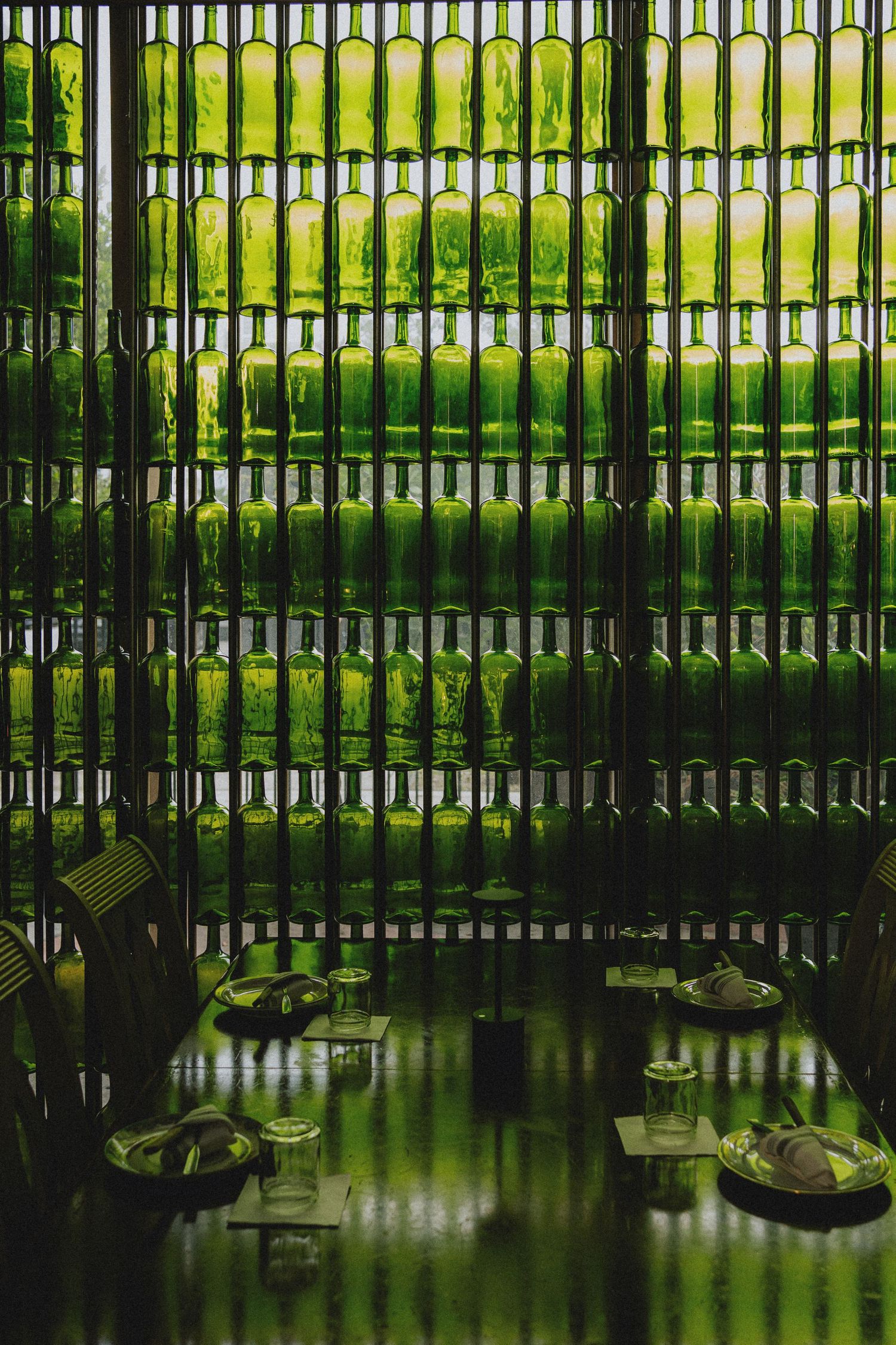 Wall display of stacked green glass bottles creating a luminous partition in a dimly lit restaurant setting.