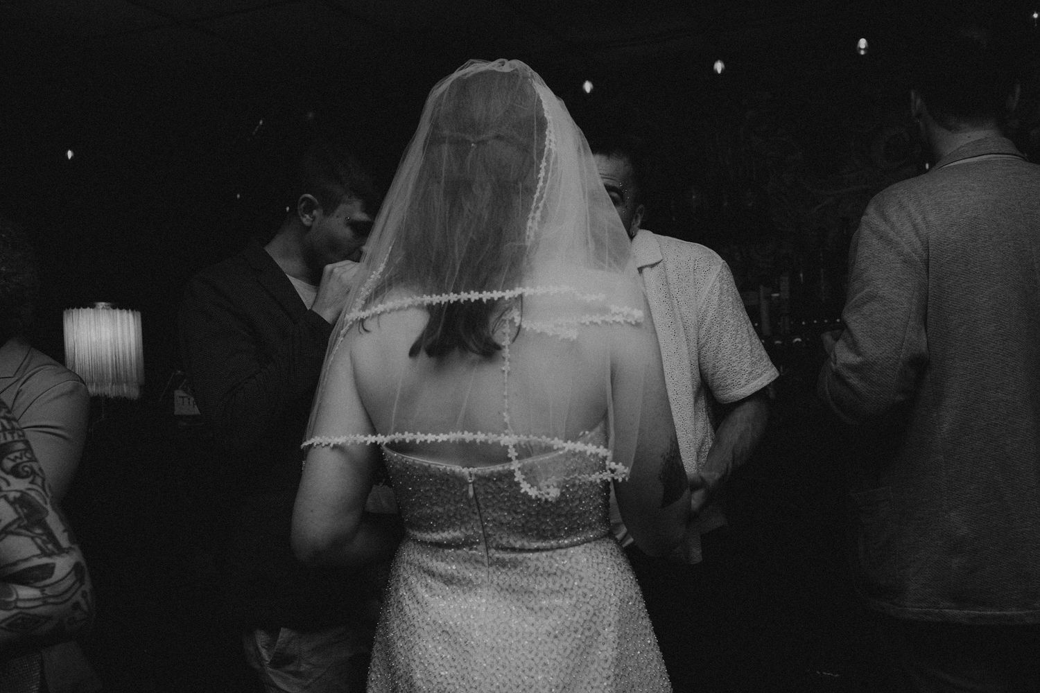 Black and white photo of a bride in a lace wedding dress and veil, seen from behind at a reception.