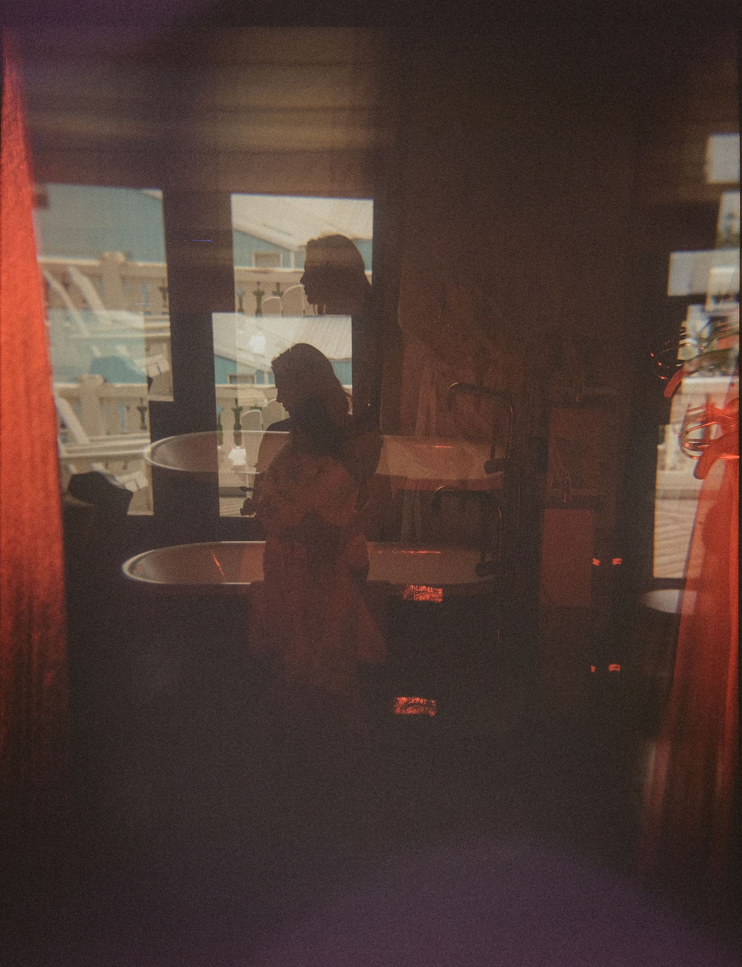 Two silhouetted figures stand near a window in a dimly lit room with warm red tones and soft ambient light.