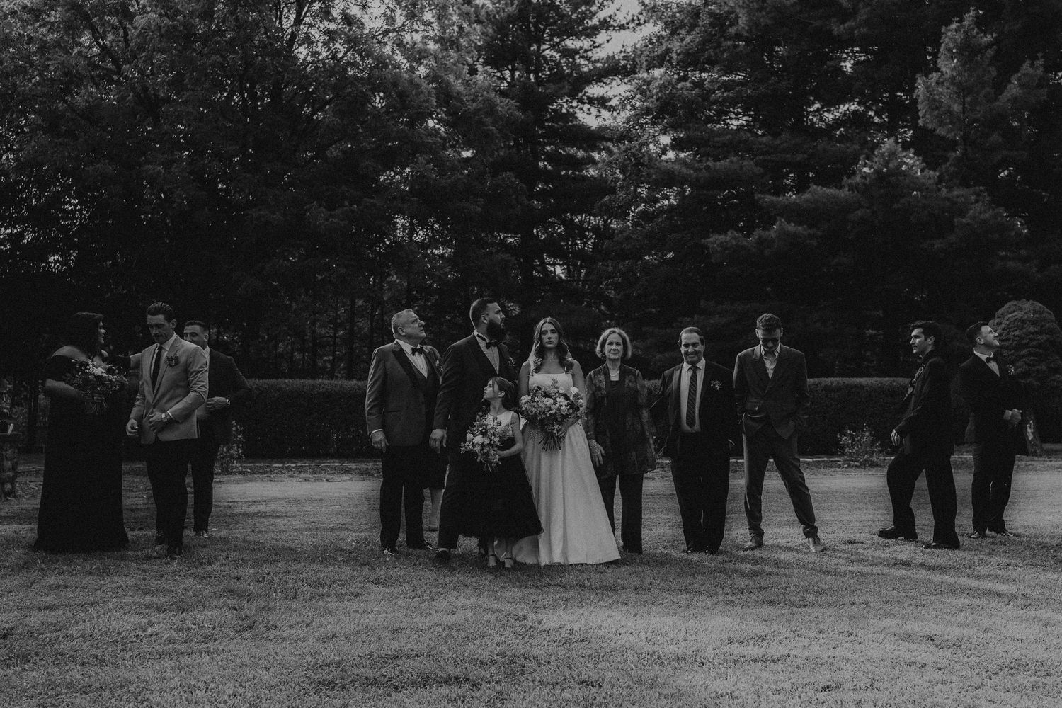 Black and white family photo outdoors, bride in white gown holding bouquet surrounded by guests near trees.