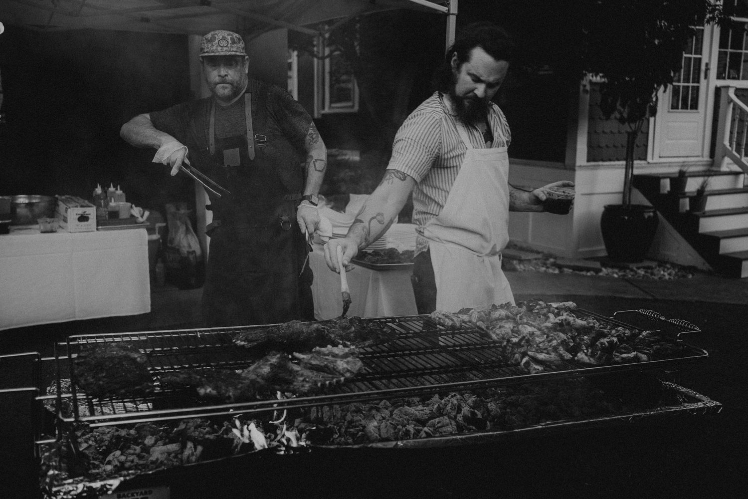 Two men grilling large cuts of meat on an outdoor BBQ grill in black and white photo.