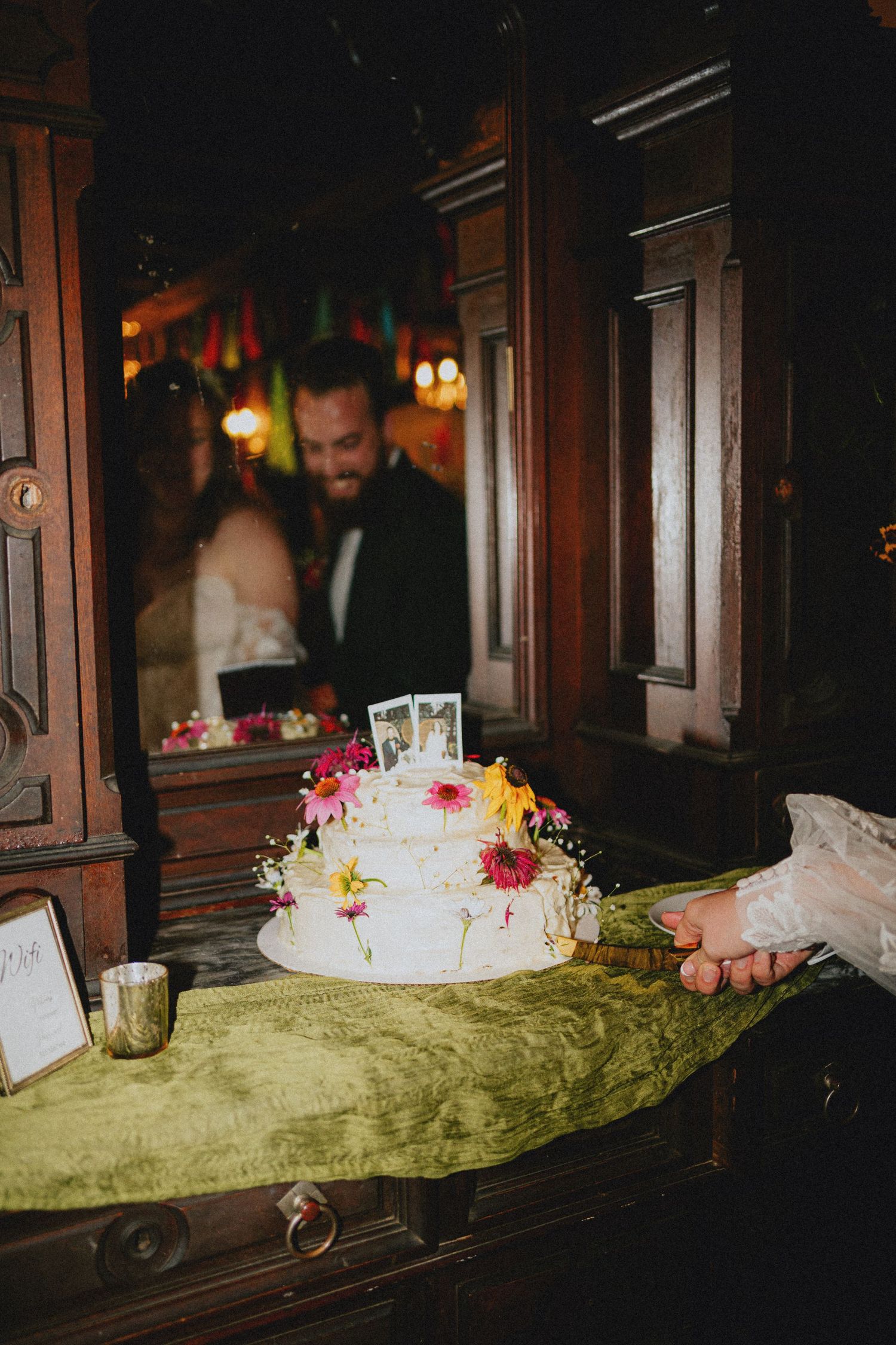 Three-tier white wedding cake adorned with colorful flowers on green velvet table, bride and groom visible in background mirror.