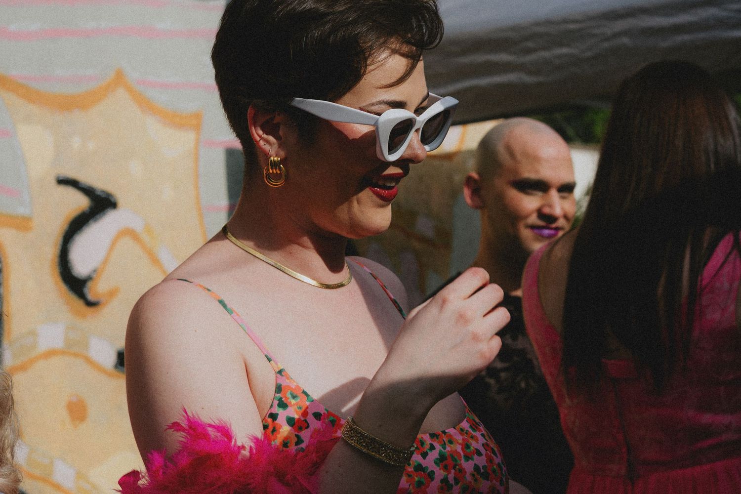 Woman in white cat-eye sunglasses and floral dress holds pink feather boa at outdoor festival event.