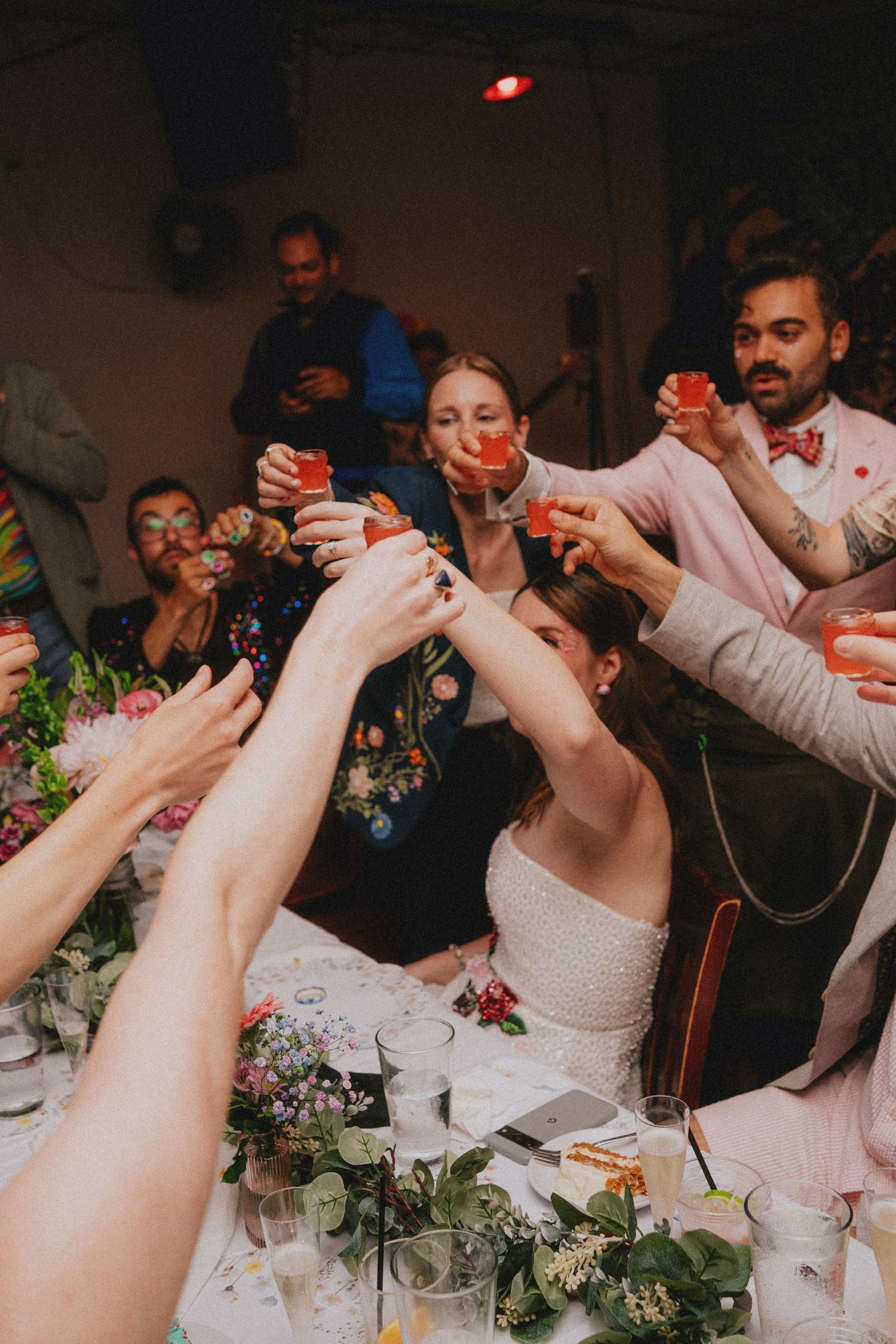 Wedding guests raising glasses for a toast at a floral-decorated reception table, with the bride in white at center.