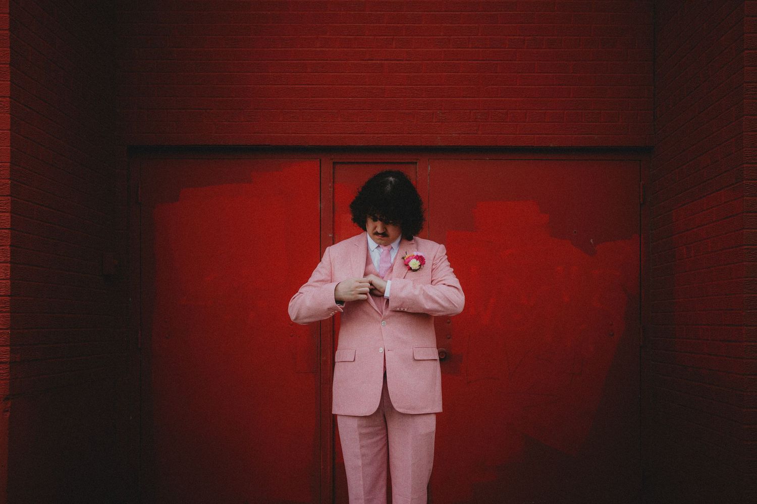 Person in pink suit with floral pin stands against vibrant red wall, looking down thoughtfully.