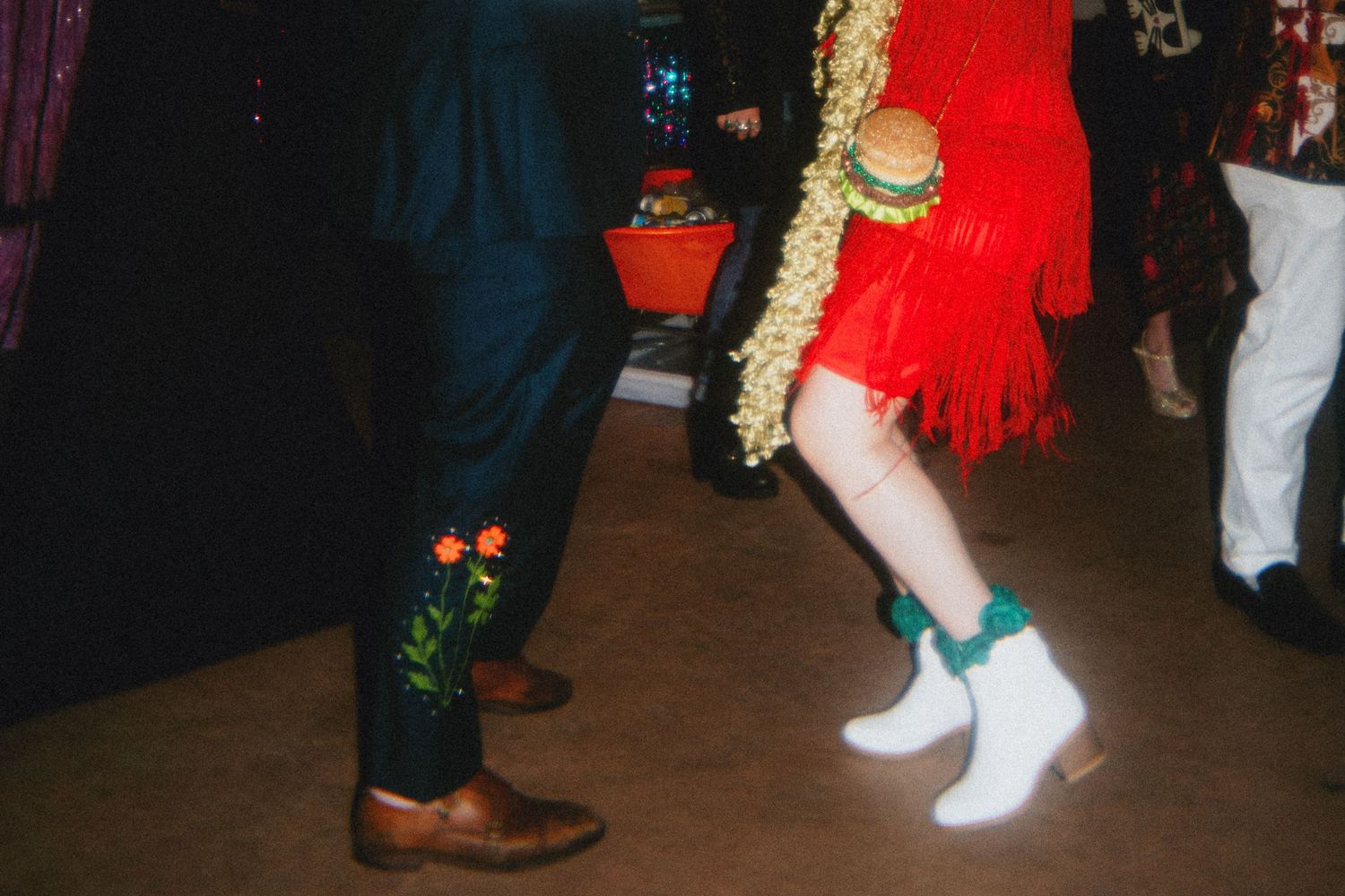Two dancers wearing cowboy boots, one in a red fringe dress with a burger purse, the other in a navy suit with floral boots.
