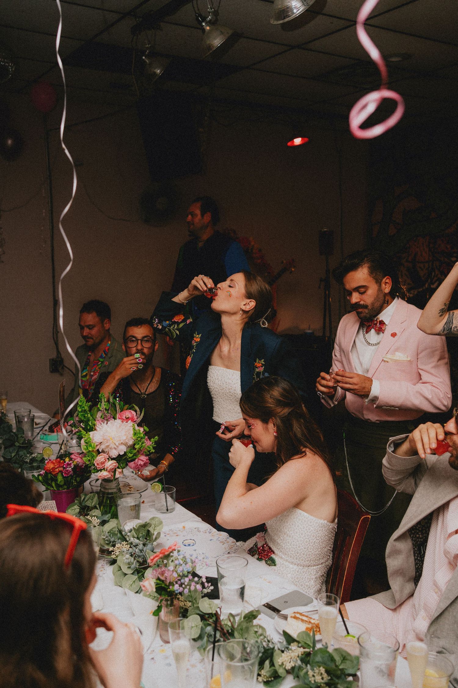 Wedding reception celebration with guests clapping, bride at head table with floral centerpieces and colorful streamers.