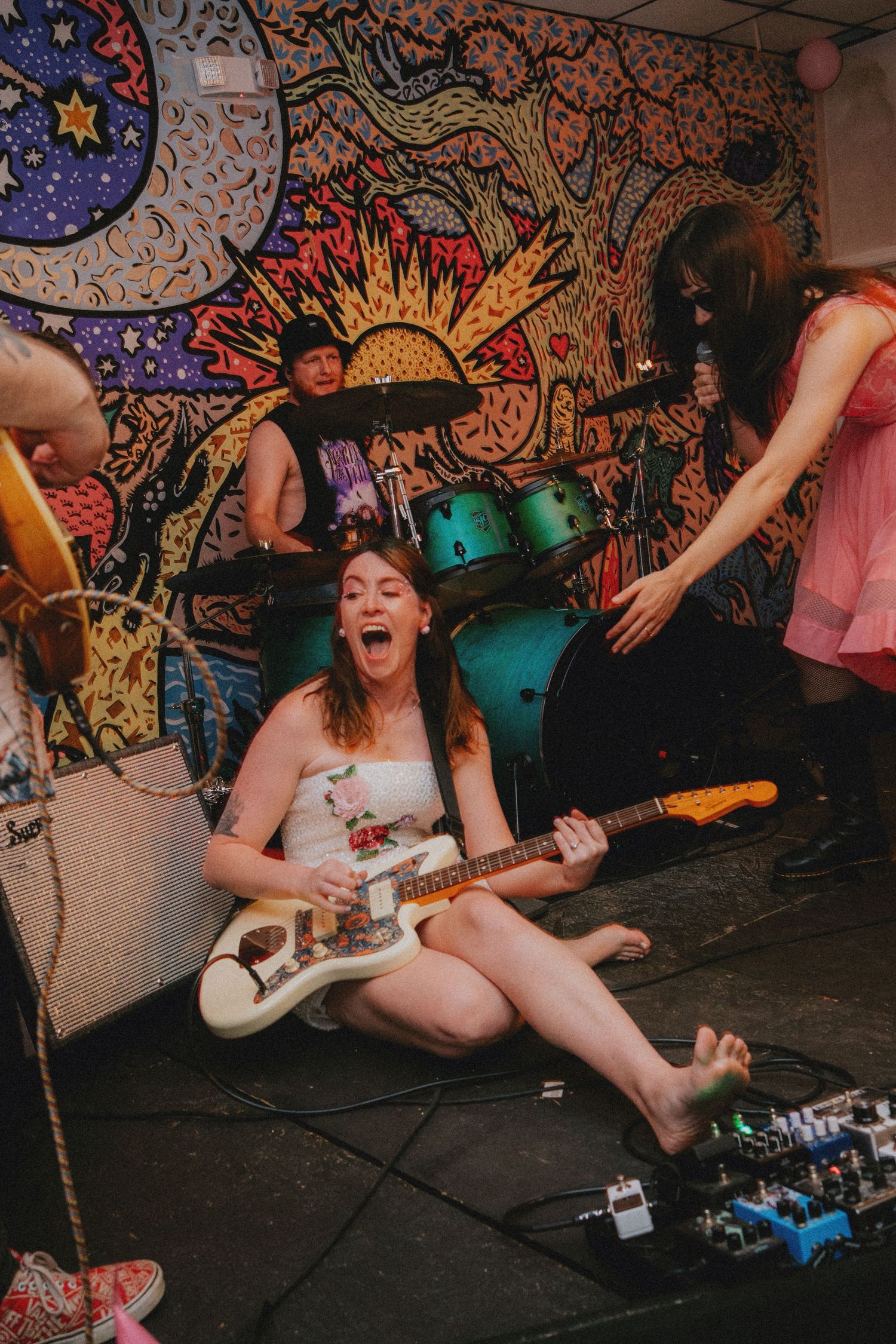 Female guitarist sits on stage floor playing white electric guitar at energetic indie rock show with colorful tapestry backdrop.