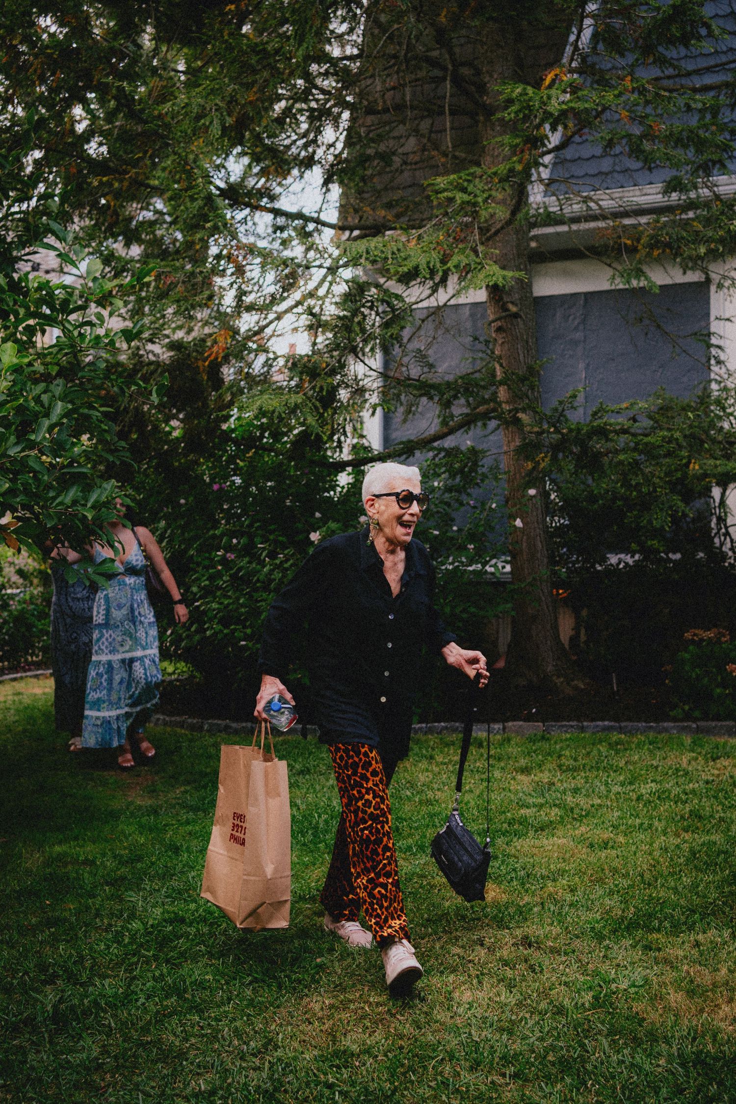 Stylish elderly woman in leopard print pants and black jacket carrying shopping bags on a green lawn.