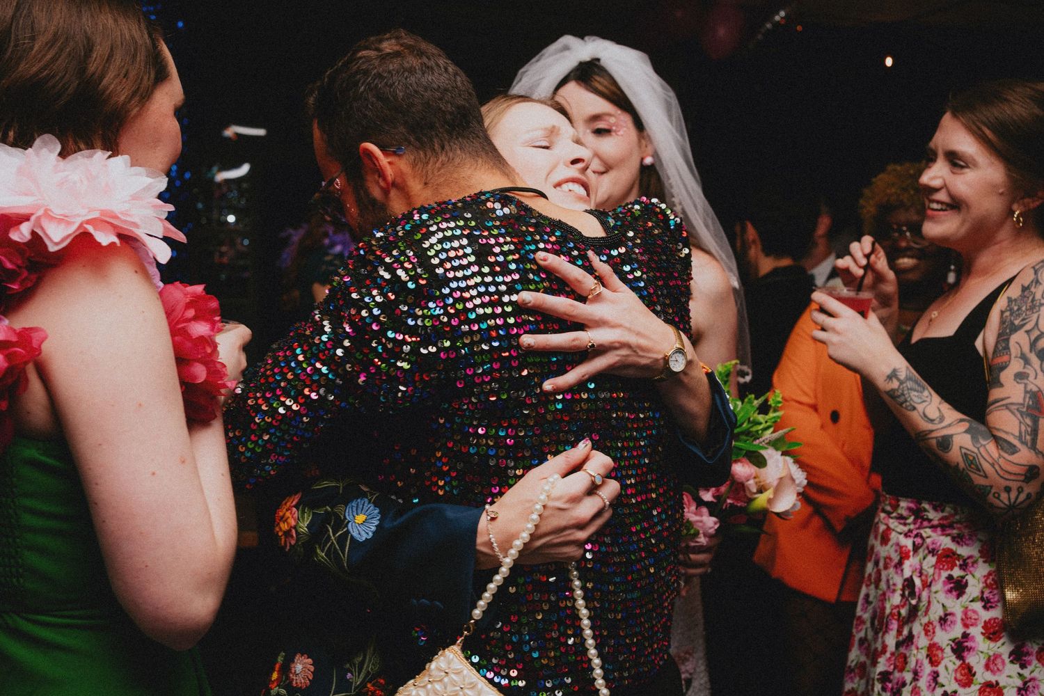 Bride in white veil hugging guest in sequin jacket on colorful wedding dance floor.