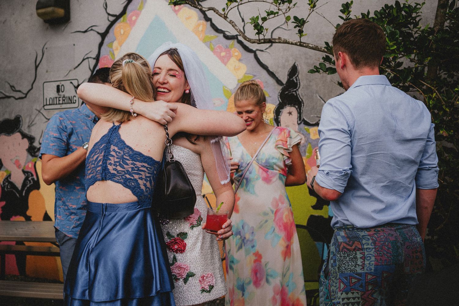 Friends hugging and celebrating at a colorful outdoor party with vibrant mural backdrop.