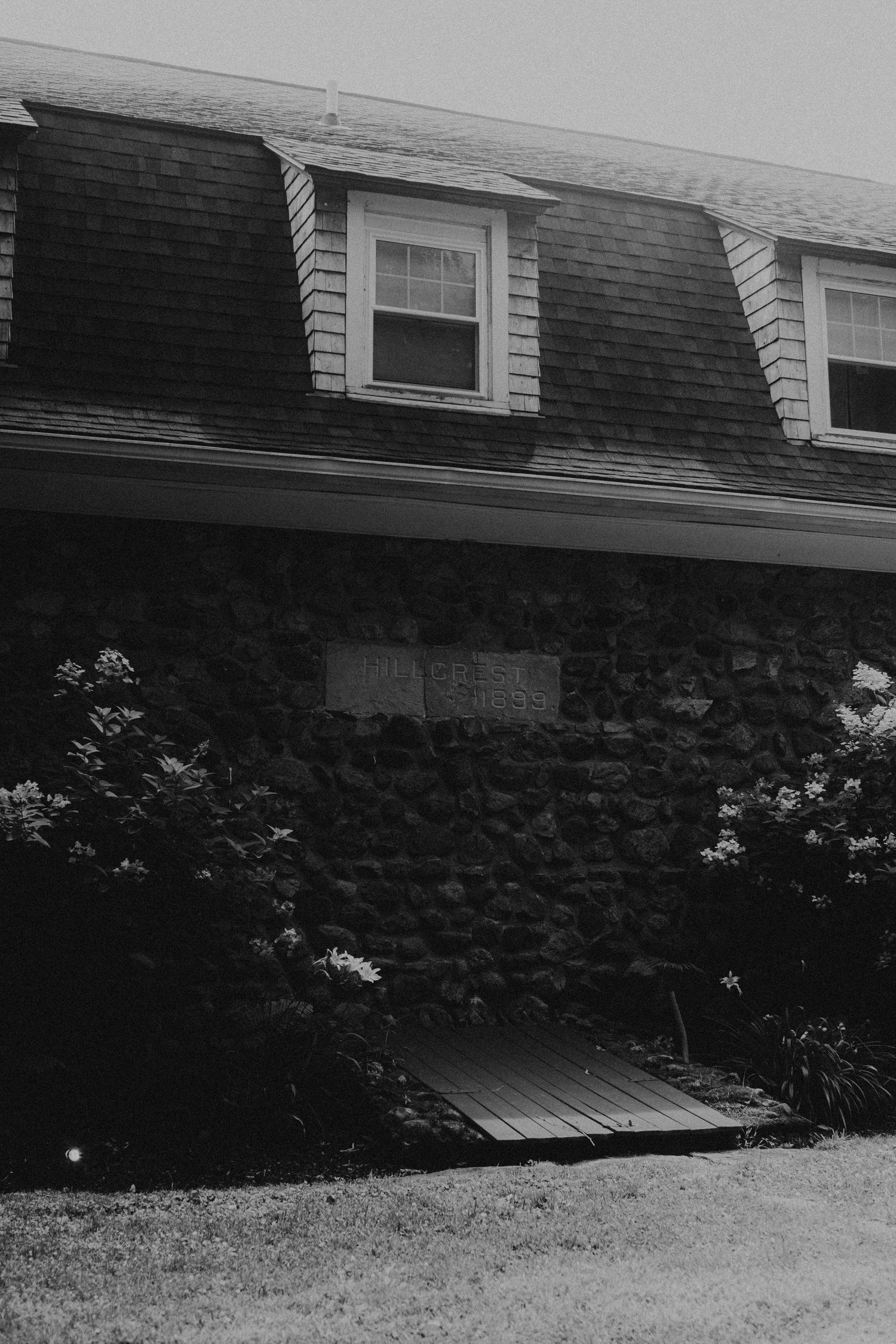Black and white photo of a stone house with dormer windows, wooden deck, and lush shrubs surrounding the exterior.