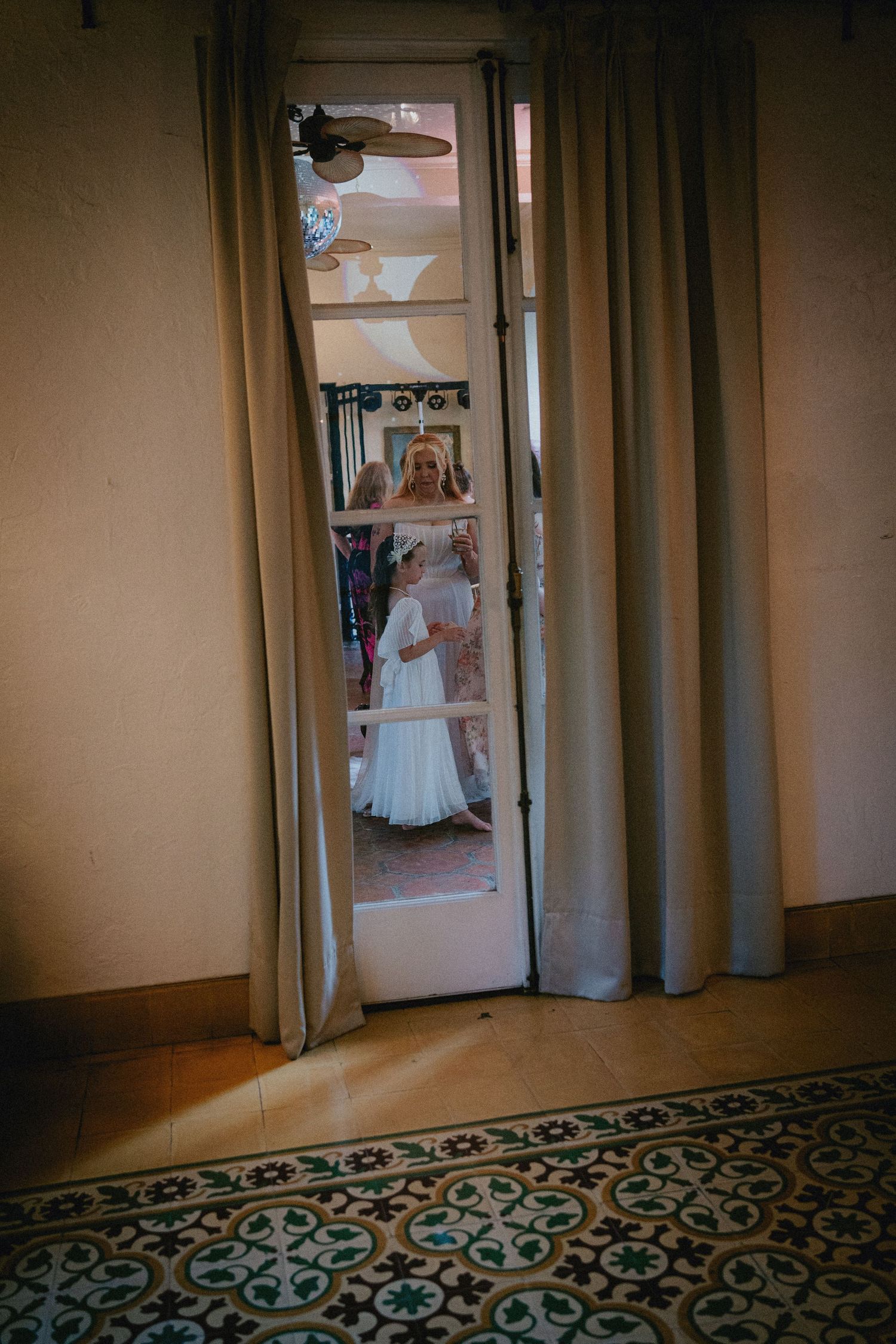Bride in white wedding dress seen through a doorway with curtains and decorative tiled floor.