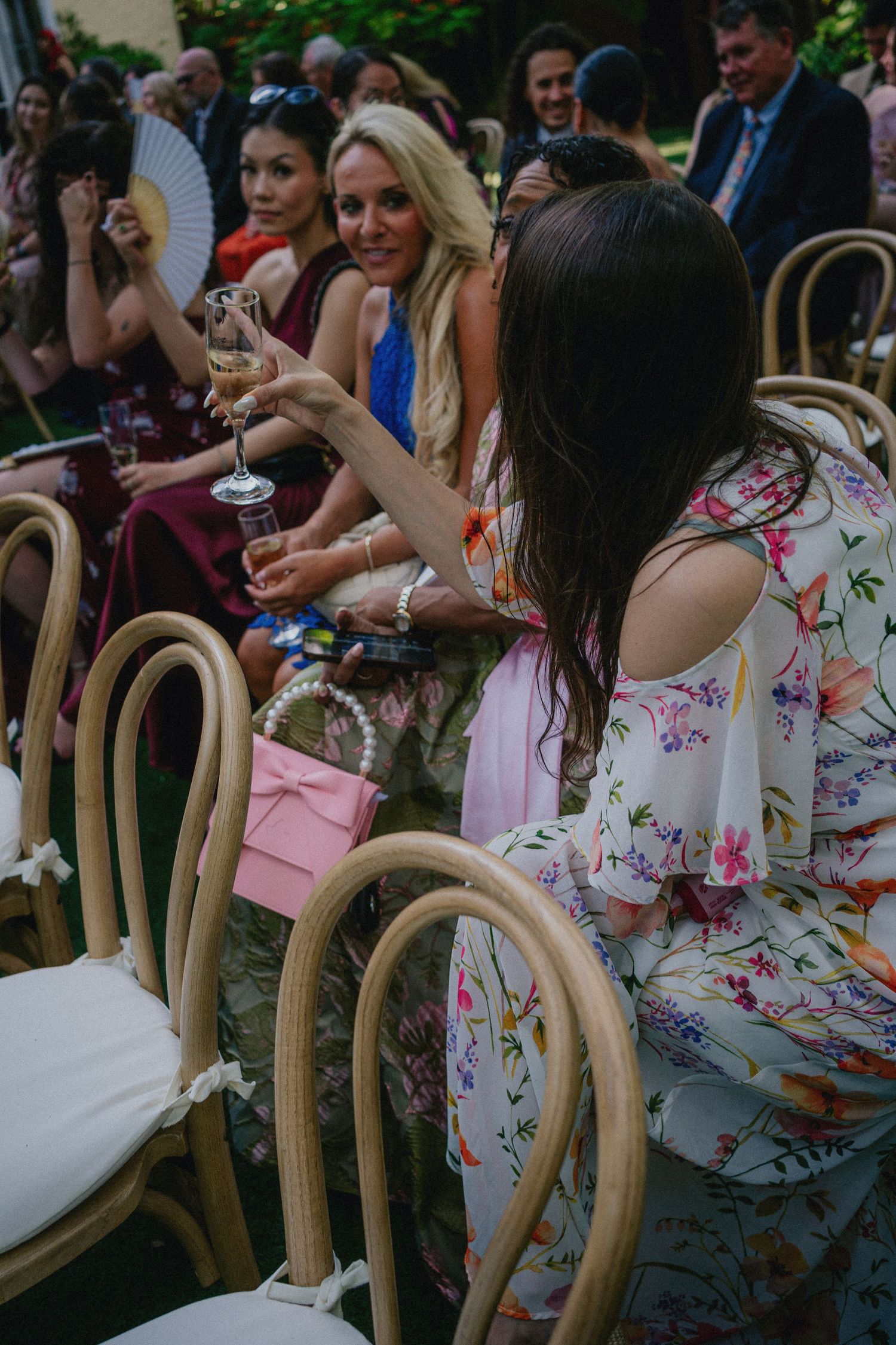 Women in floral dresses toasting with champagne glasses at an outdoor wedding reception seated on bentwood chairs.