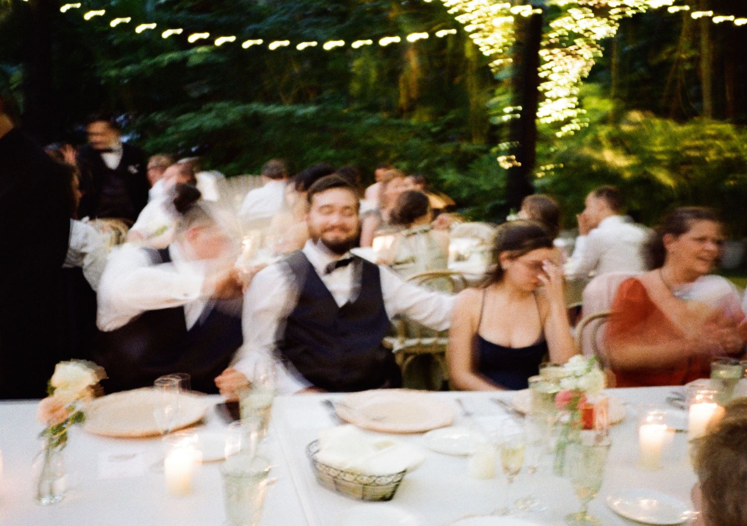 Guests laughing and celebrating at an outdoor wedding reception dinner with candles and string lights.