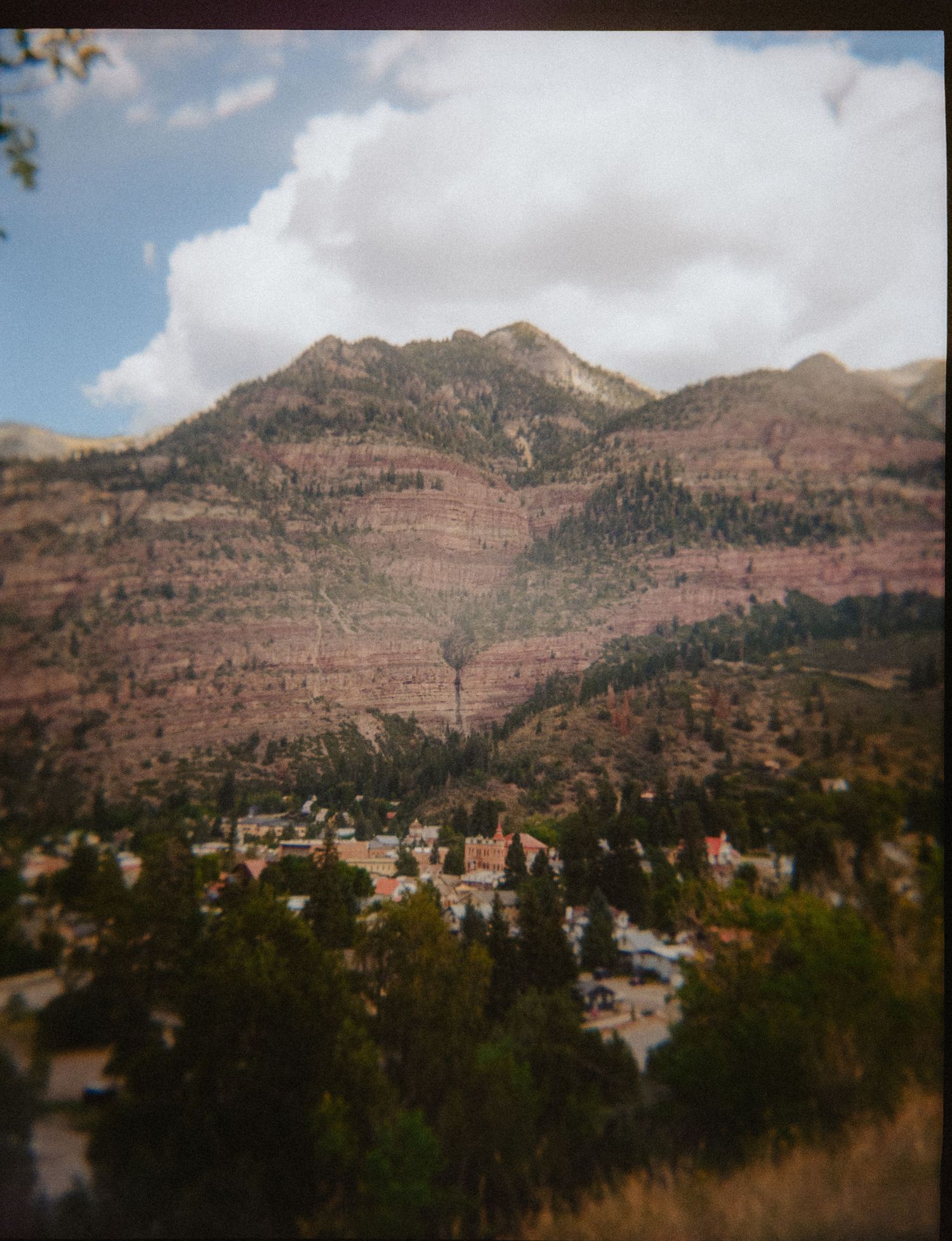 Aerial view of a small mountain town nestled among trees with dramatic red rock mountains and clouds in the background.