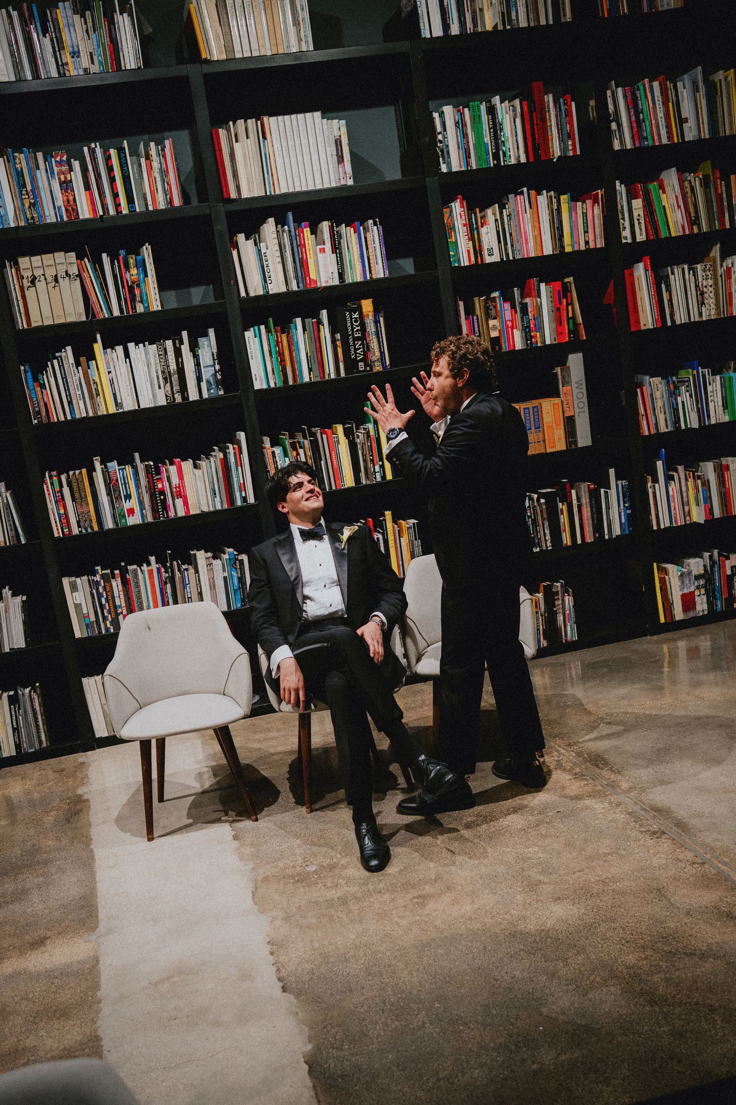 Two men in tuxedos converse in a library with floor-to-ceiling bookshelves, one seated and one standing gesturing.