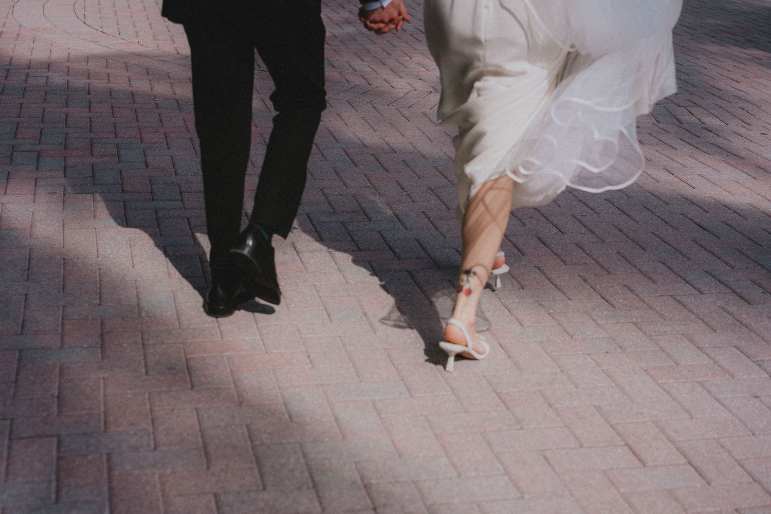 Bride and groom walking on brick pavement, showing wedding dress hem and heels beside groom's black dress shoes.