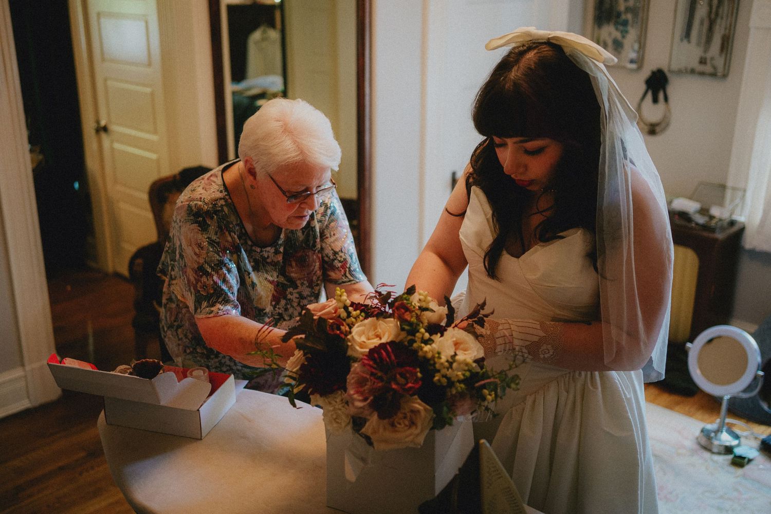 Elderly woman helping bride with floral bouquet in elegant room, bride wearing white gown and veil on wedding day.