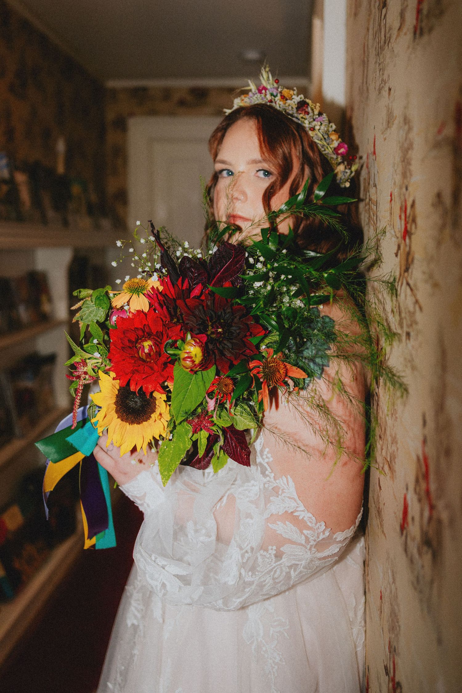 Bride in lace gown holding vibrant sunflower bouquet with floral crown, posing against floral wallpaper.