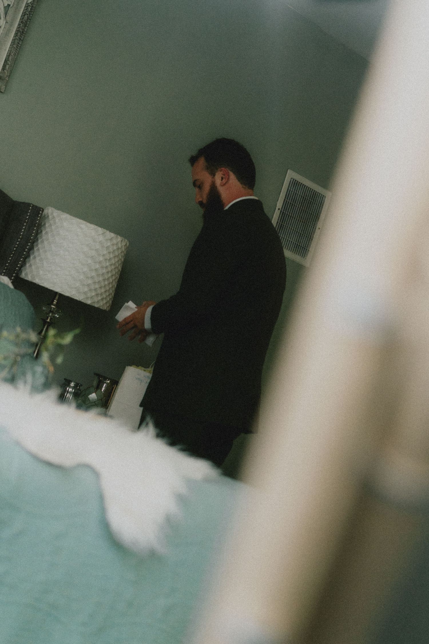 A bearded groom in a black suit prepares in a softly lit room, reflected in a mirror on his wedding day.