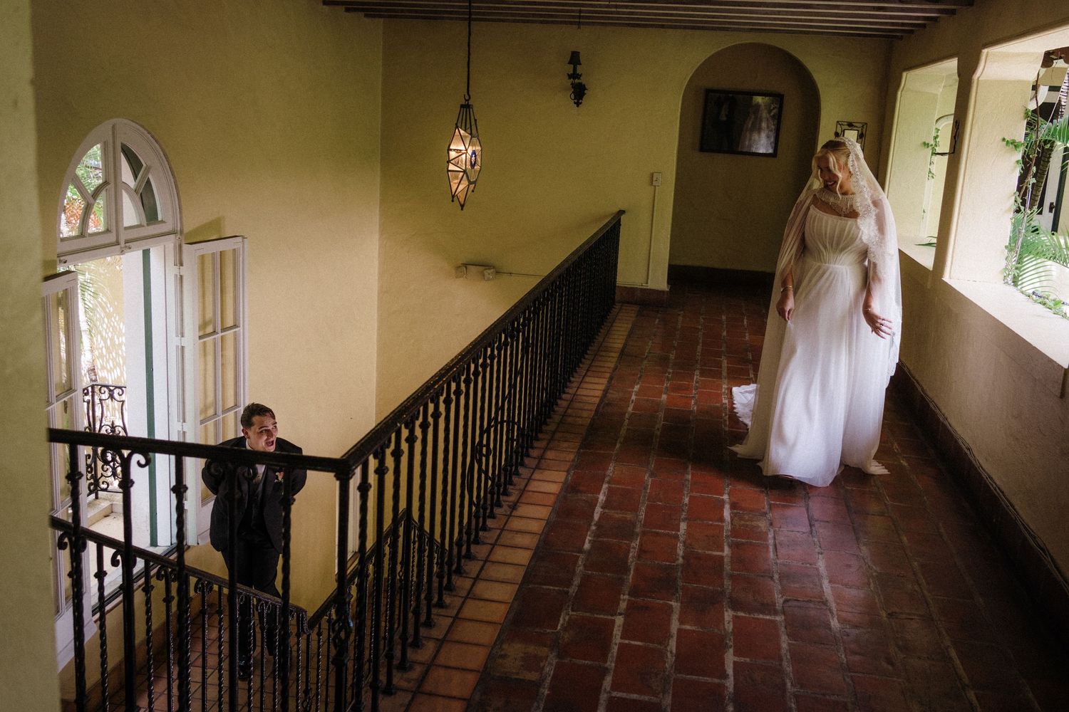 Bride in white gown stands on terracotta-tiled hallway balcony as young boy in suit watches near ornate staircase railing.