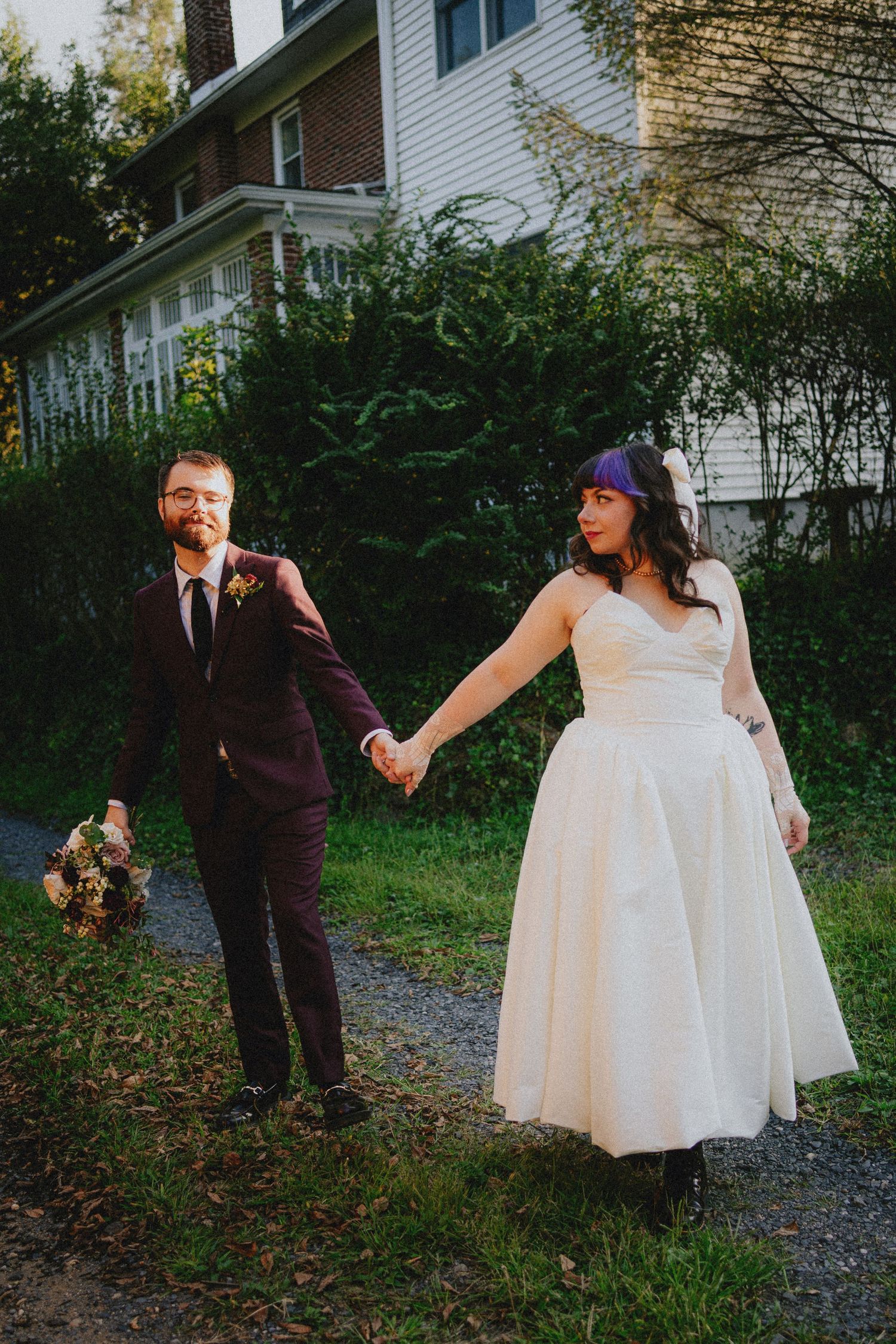 A bride in a white tea-length dress and groom in a burgundy suit hold hands while walking outdoors on their wedding day.