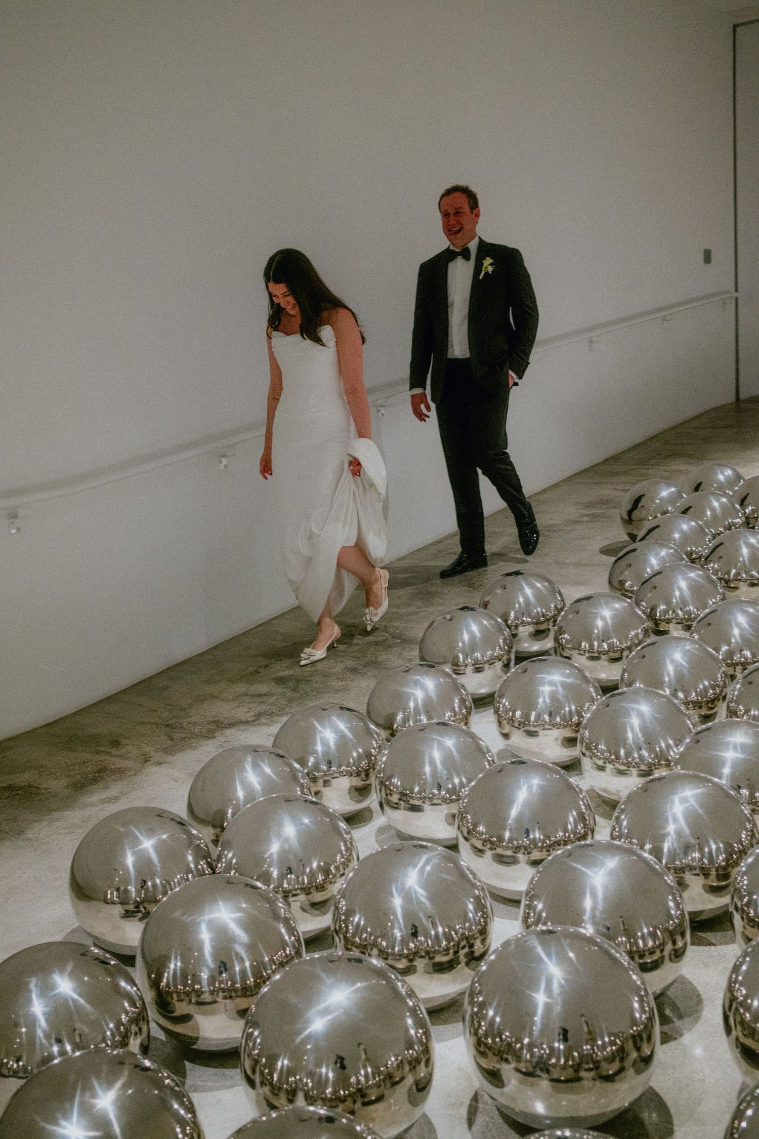 Bride and groom walking among shiny silver mirrored spheres on a gallery floor during their wedding.