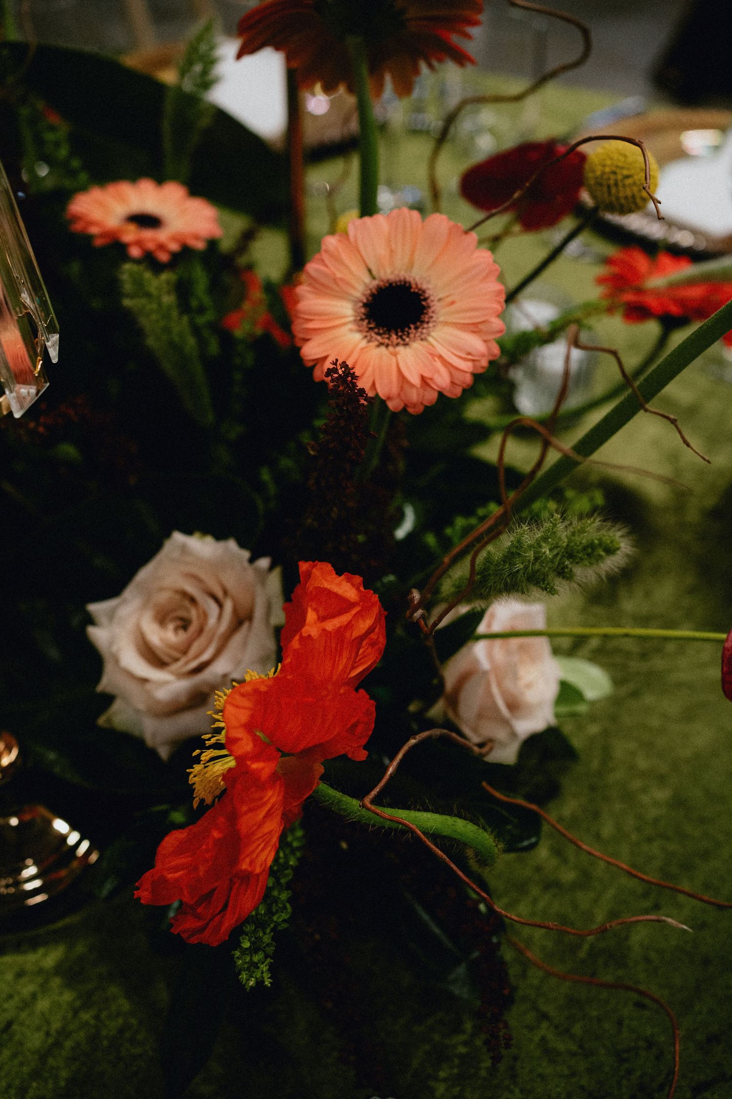 Elegant floral centerpiece with coral gerbera daisies, red poppies, and cream roses on a lush green moss table.