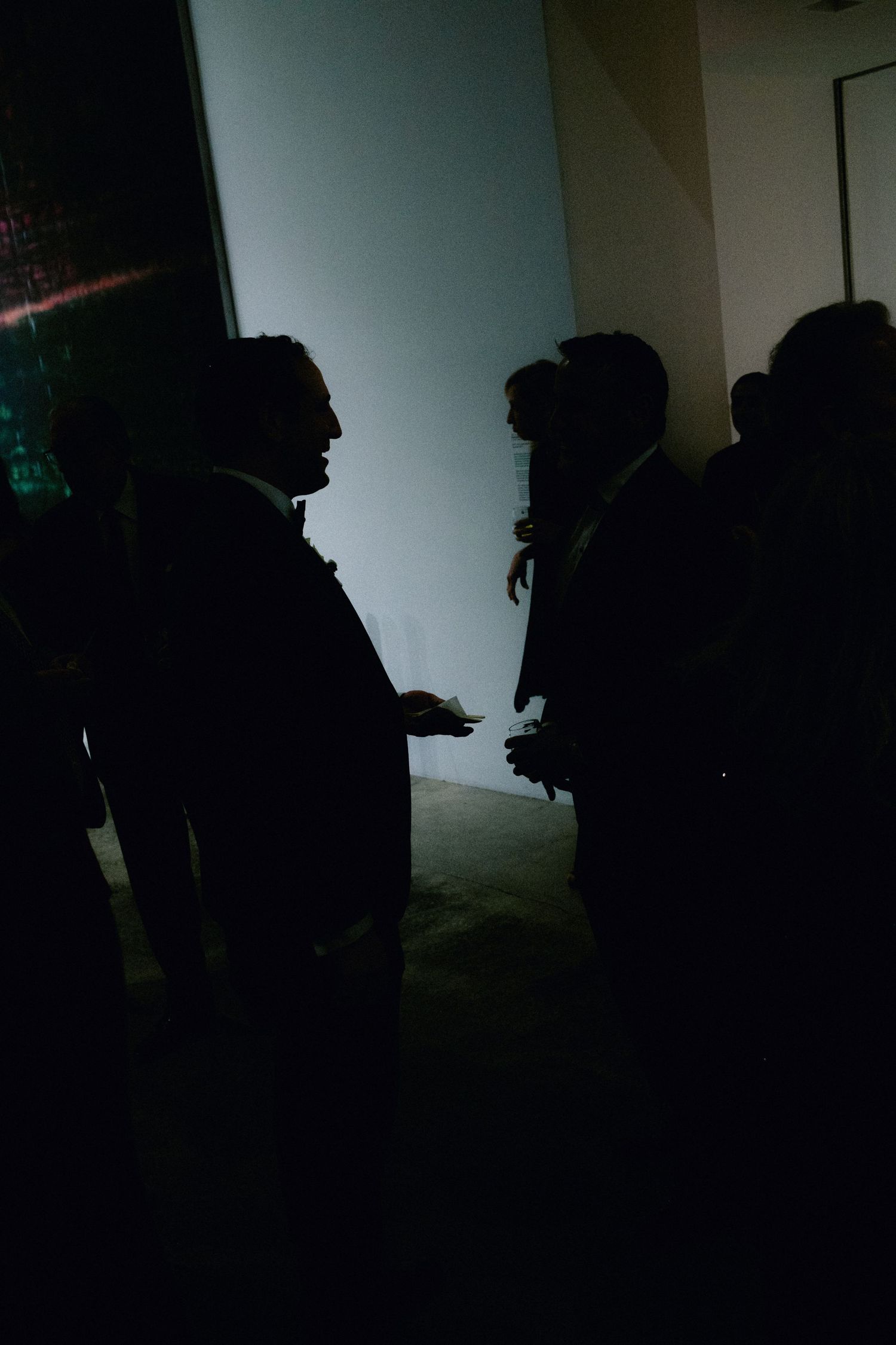 Silhouettes of people in formal attire conversing in a dimly lit room with a glowing white wall backdrop.