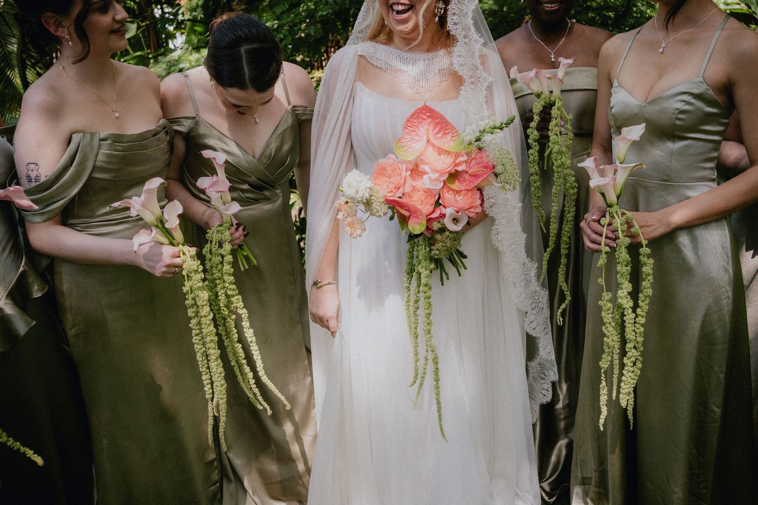 Bride in white gown with veil laughs holding coral bouquet, flanked by bridesmaids in olive satin dresses with green trailing bouquets.