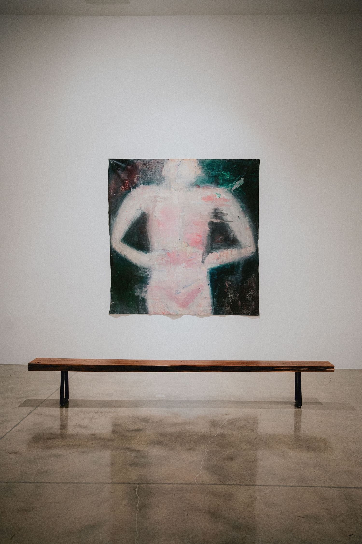 A wooden bench sits before an abstract painting of a human figure with visible spine on a white gallery wall.