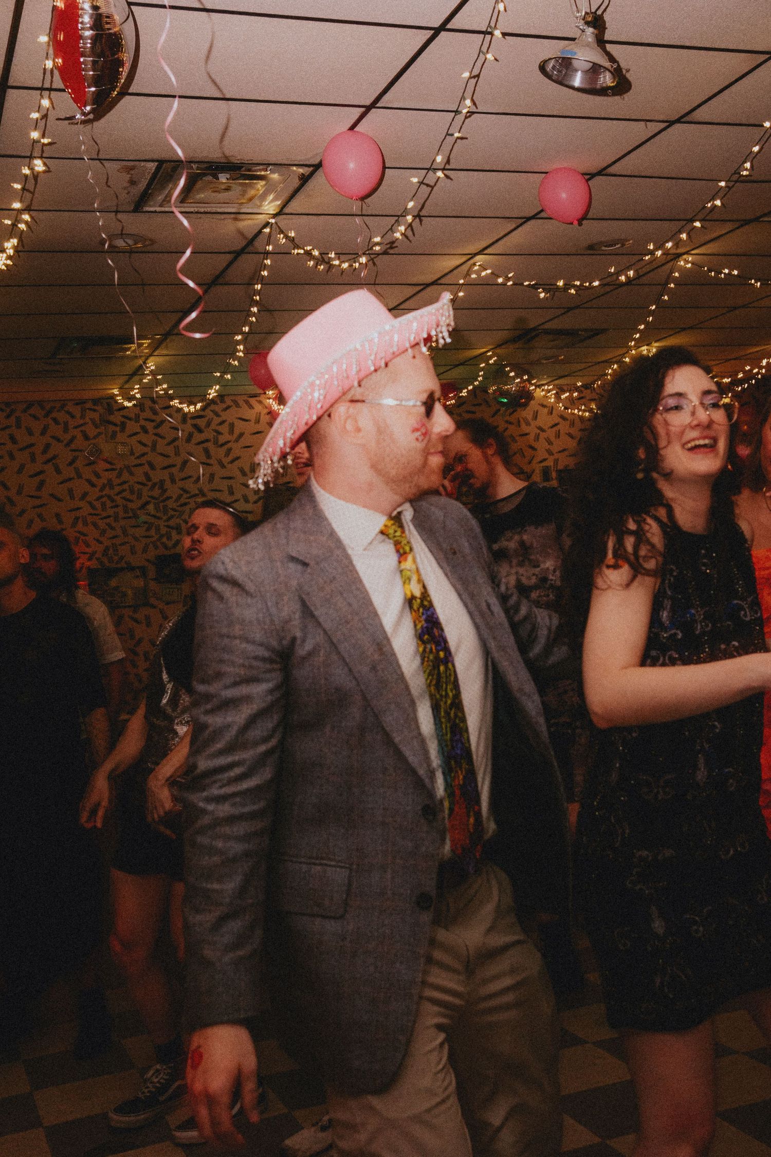 Man in pink fringed hat and gray blazer dancing at a festive party with balloons and string lights.