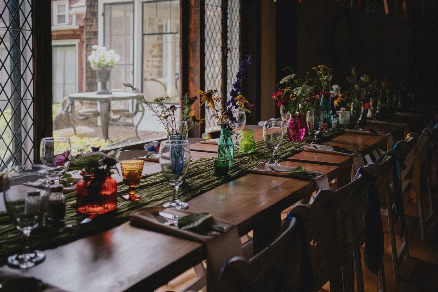 Elegant long wooden dining table set with colorful glass vases, flowers, and place settings in a rustic venue with leaded windows.