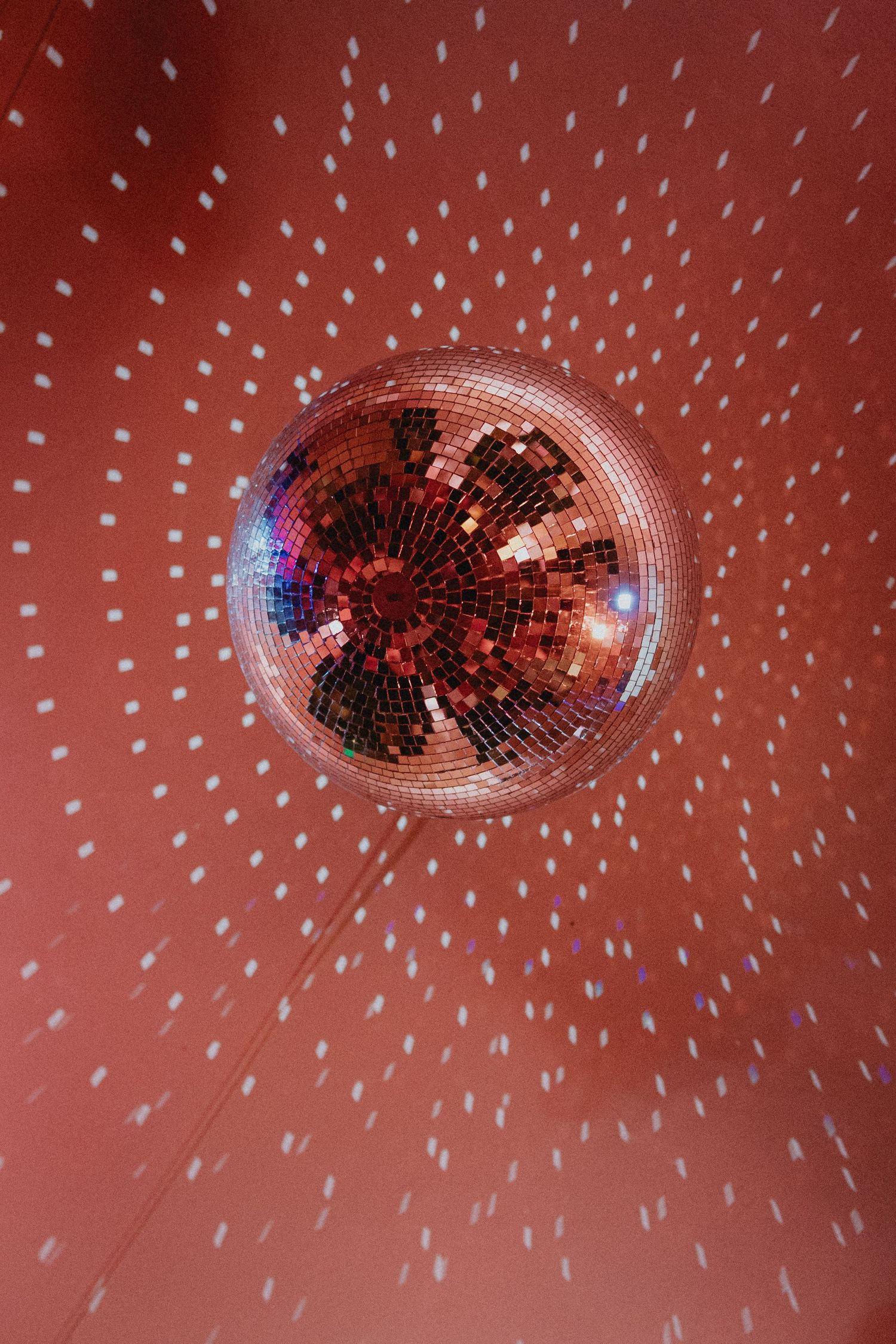 A shimmering disco ball reflecting red and white light patterns on a coral-colored ceiling in a festive party setting.