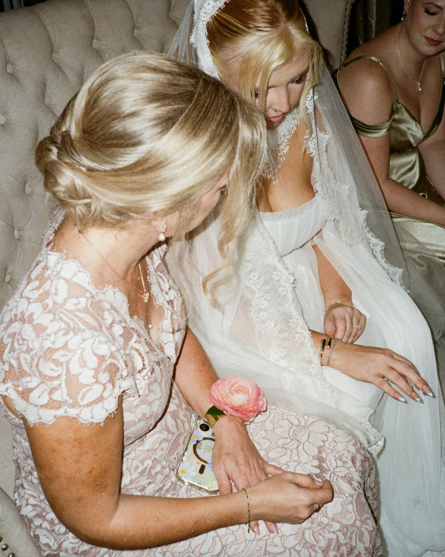 Bride in white lace gown and veil sits with blonde woman in pink lace dress adjusting her wrist corsage.