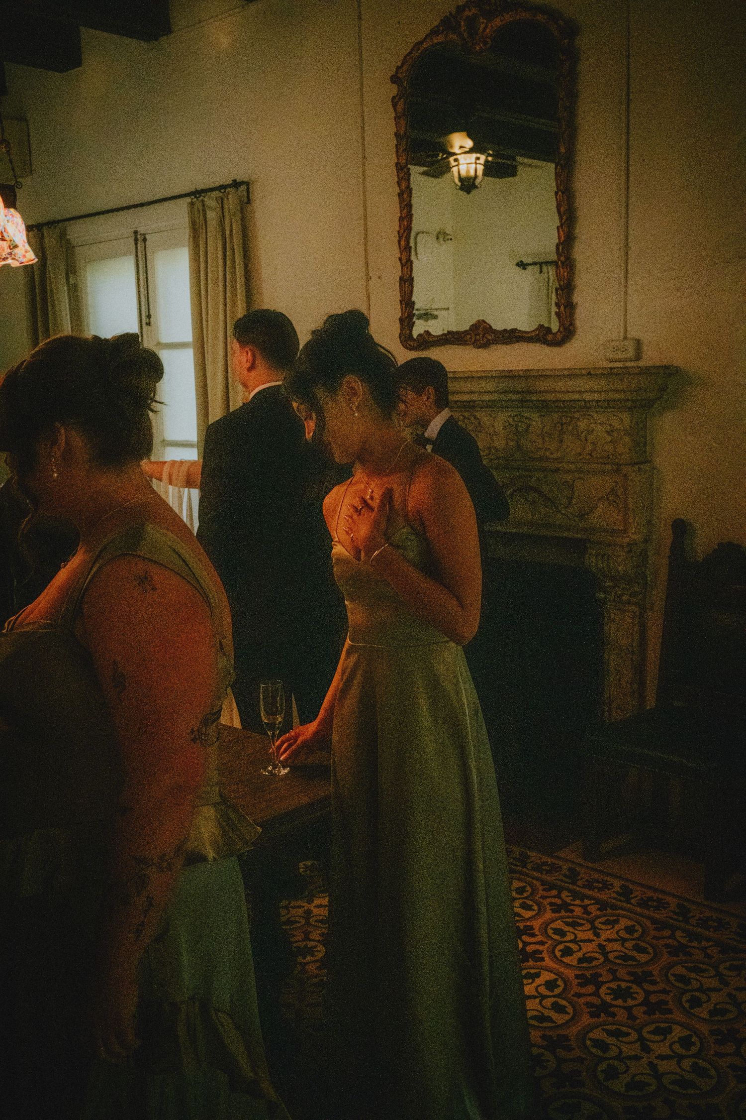 Elegant guests mingle in a warmly lit vintage room with ornate fireplace and mirror during a formal evening event.